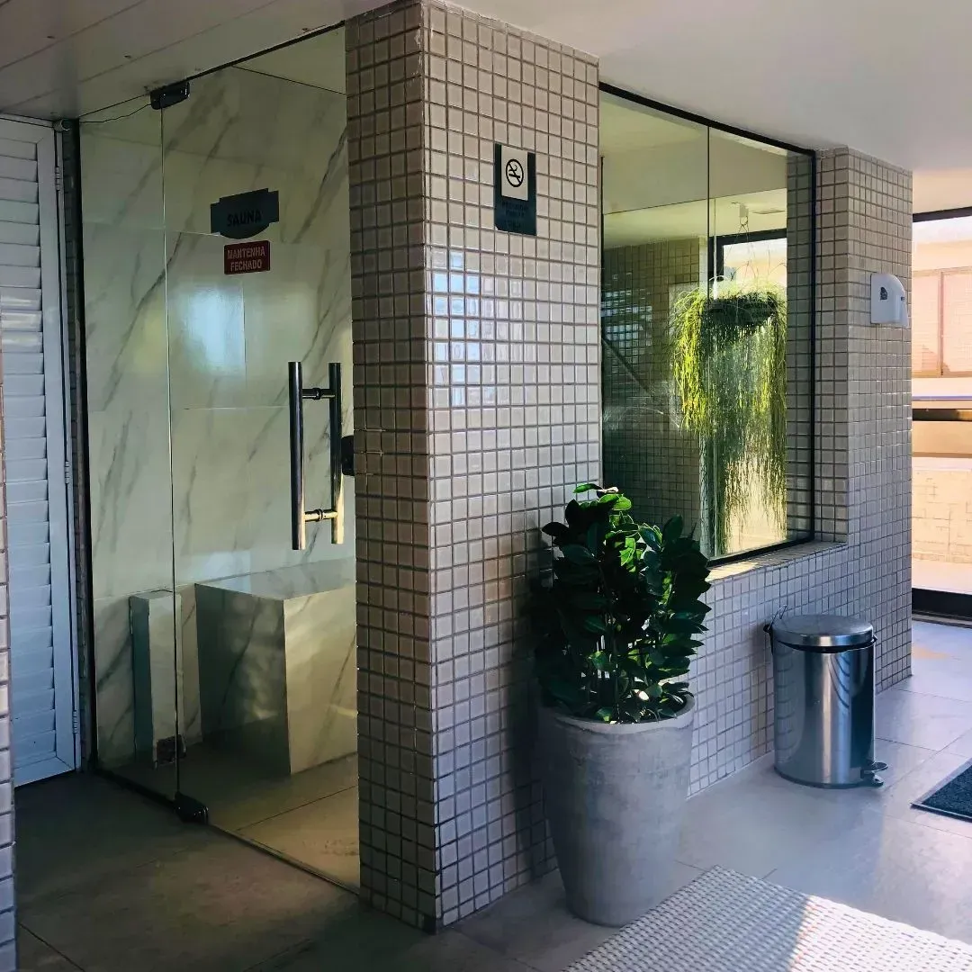 Entrada para uma sauna com portas de vidro, paredes revestidas com azulejos brancos e um vaso de planta.