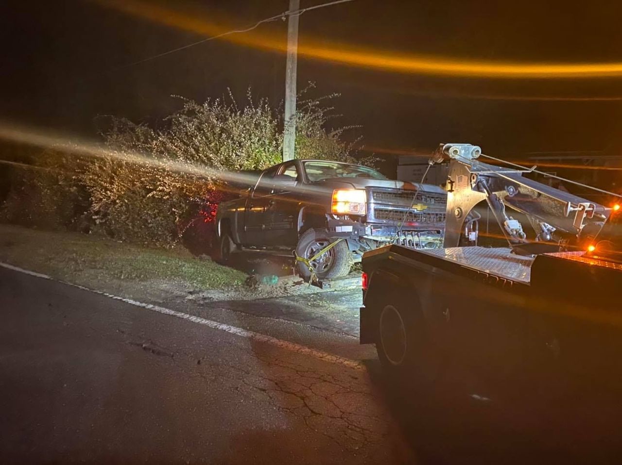 A tow truck pulls a damaged black truck from roadside bushes at night.