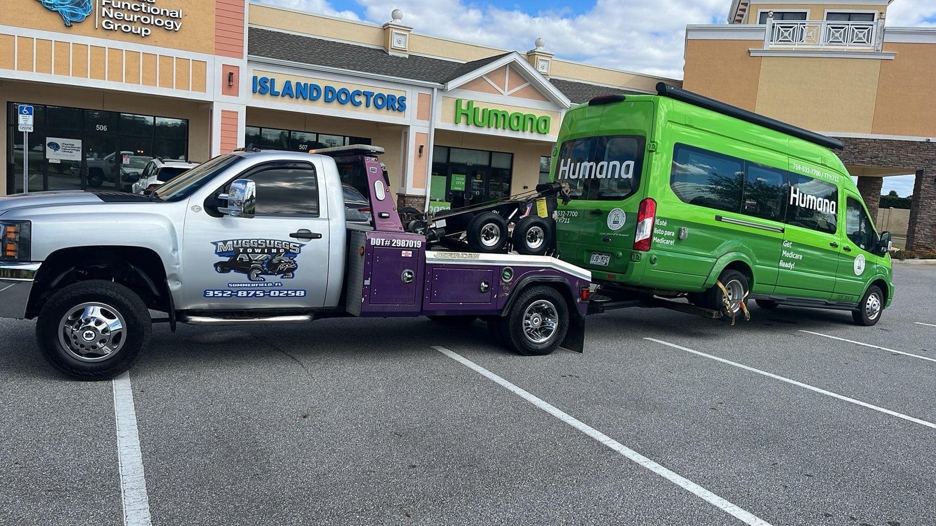 Tow truck towing a green shuttle bus in a parking lot, near a shopping center.