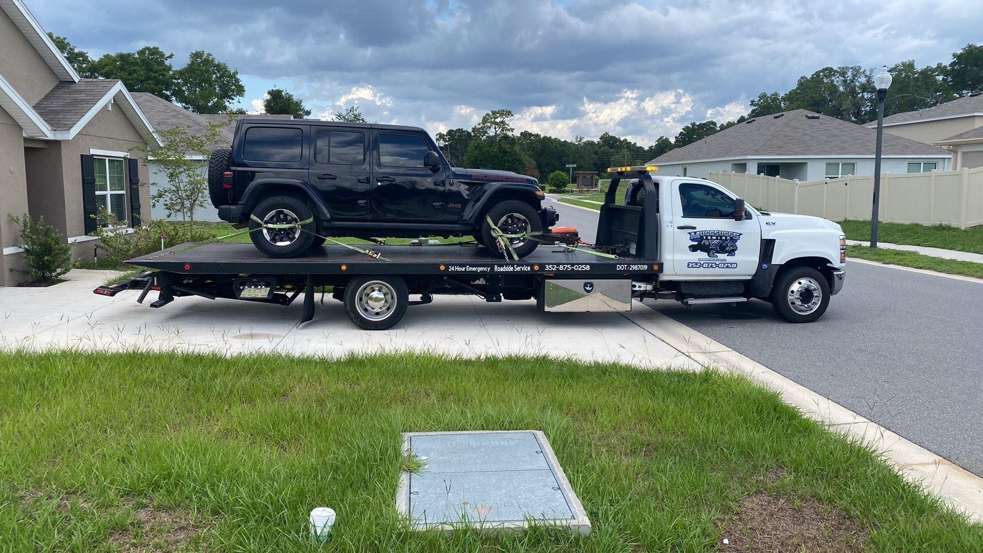 Black Jeep being transported on a flatbed tow truck in a residential area. Overcast sky.