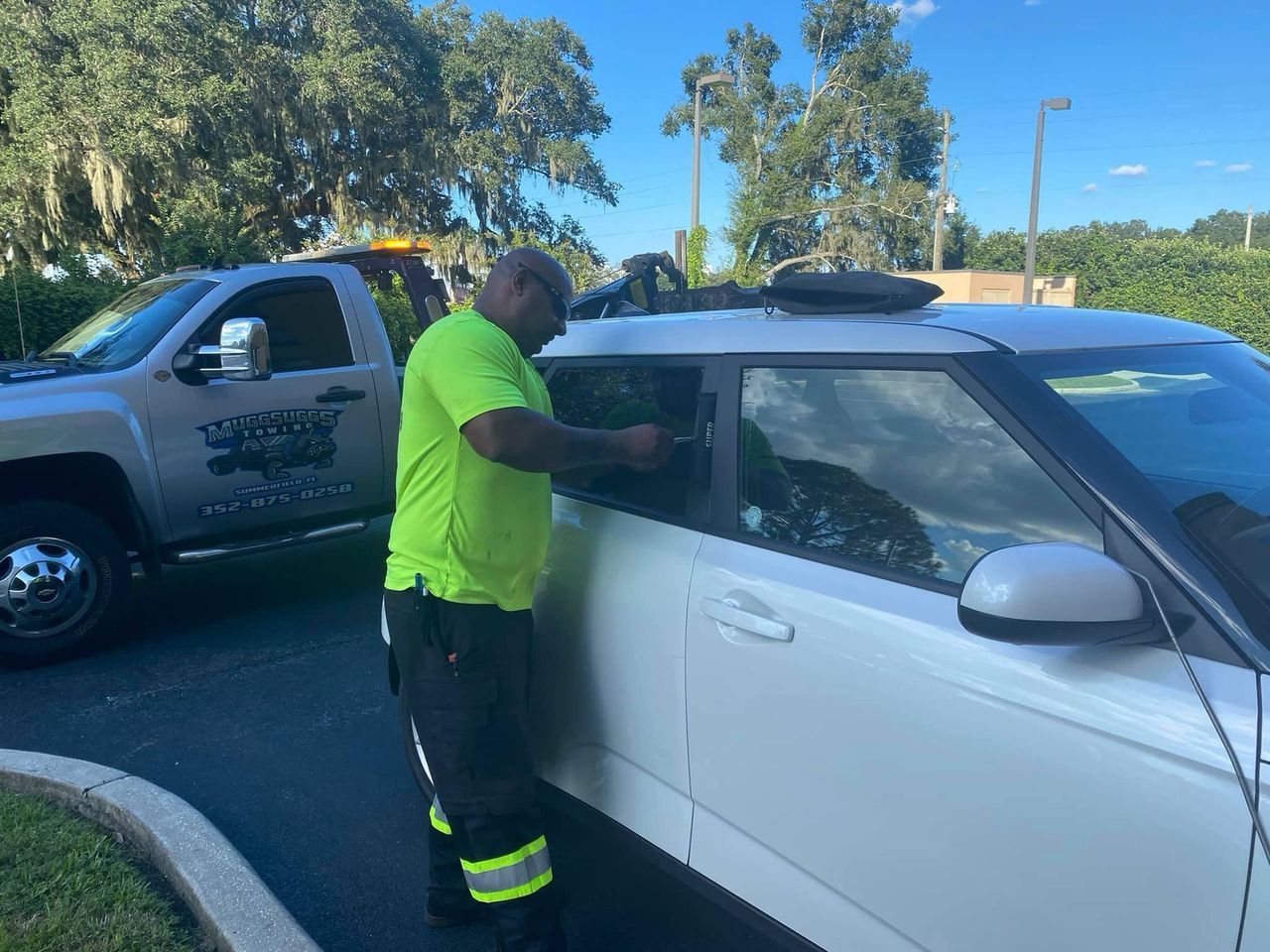 A man in neon green unlocks a white car. A tow truck is parked nearby. Outdoors.