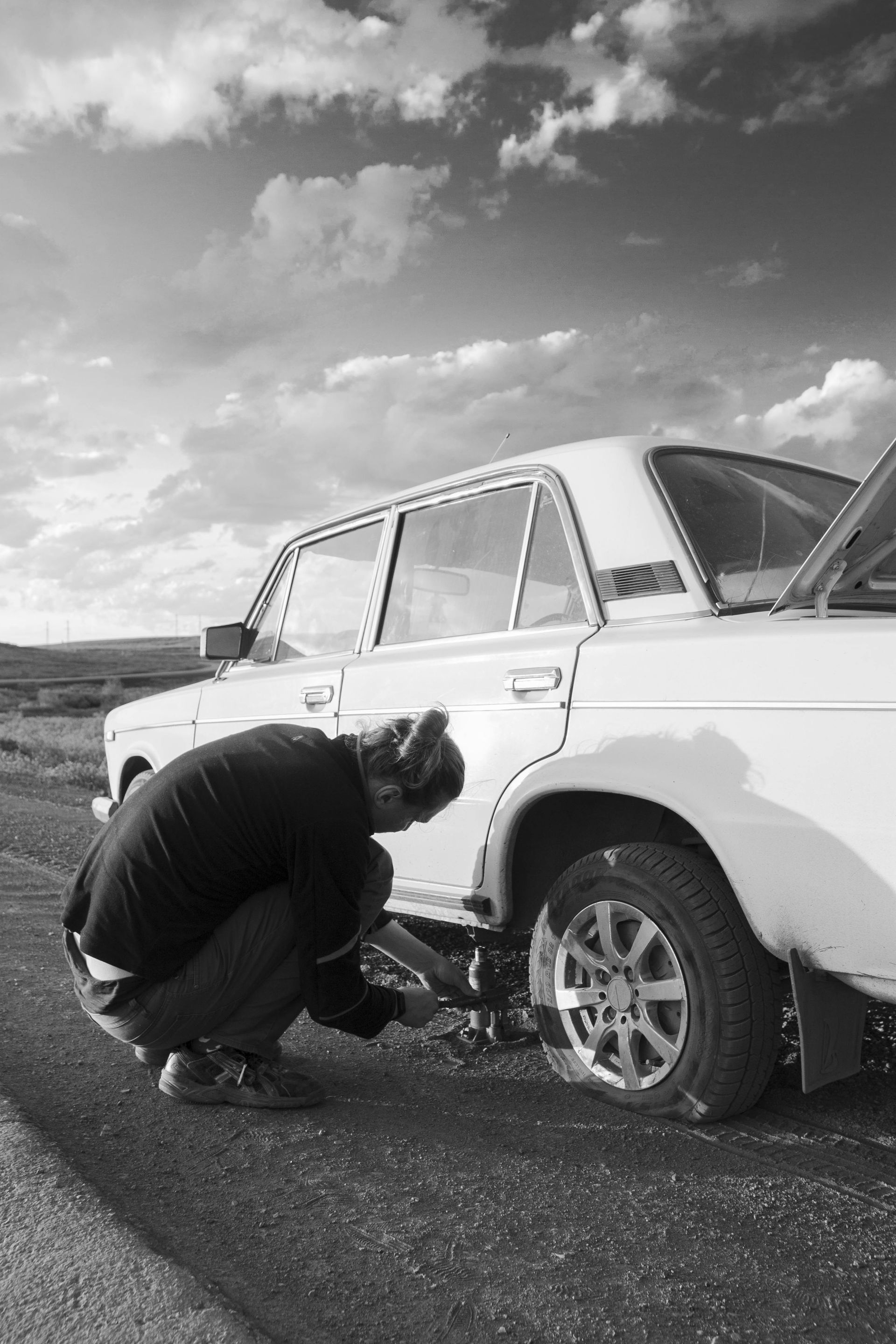 Person changing a flat tire on a white car on the side of a road, with a cloudy sky.