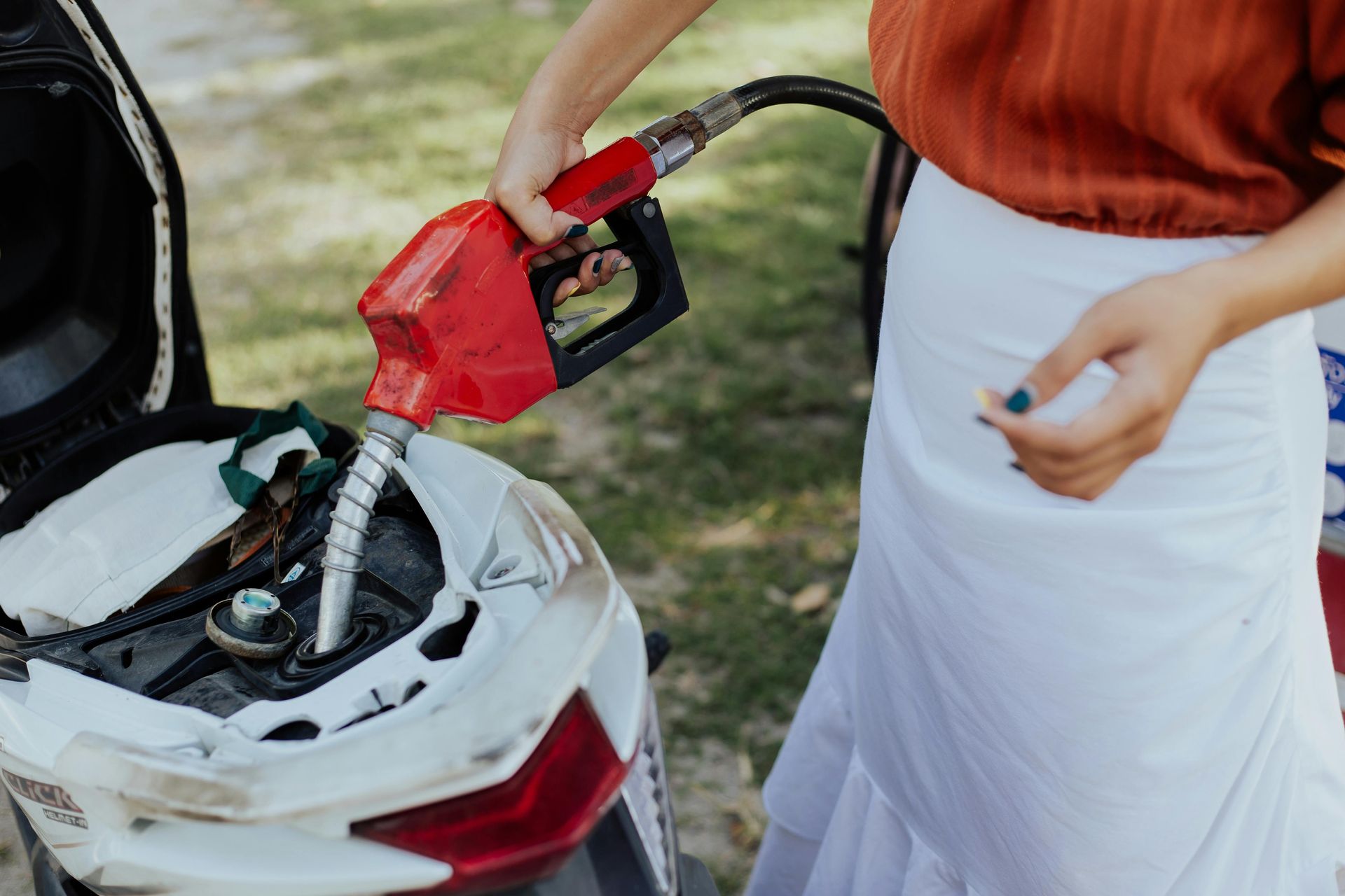 Person filling a scooter's gas tank with a red fuel pump outdoors.