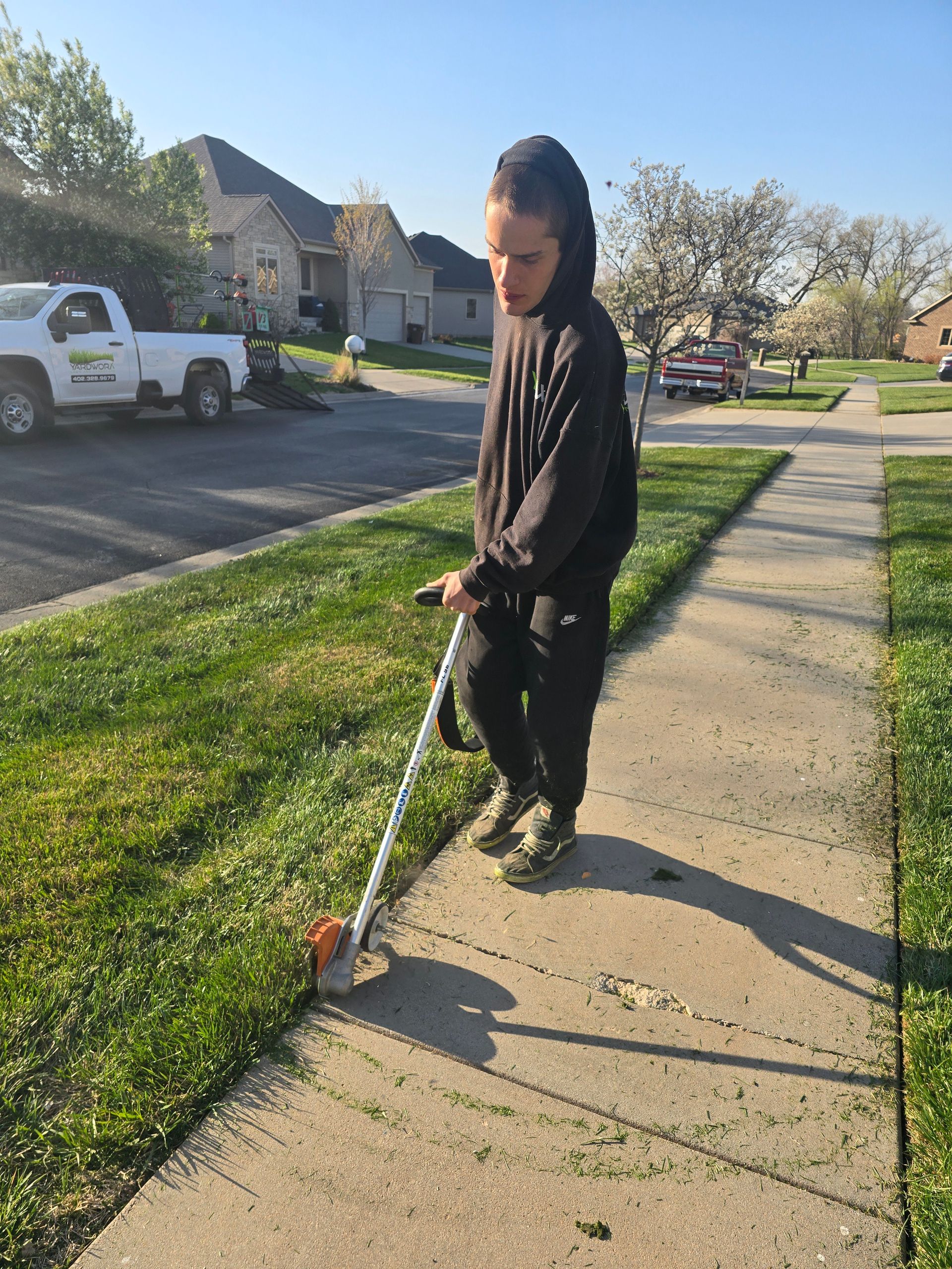 A man is standing on a sidewalk cutting grass with a lawn mower.