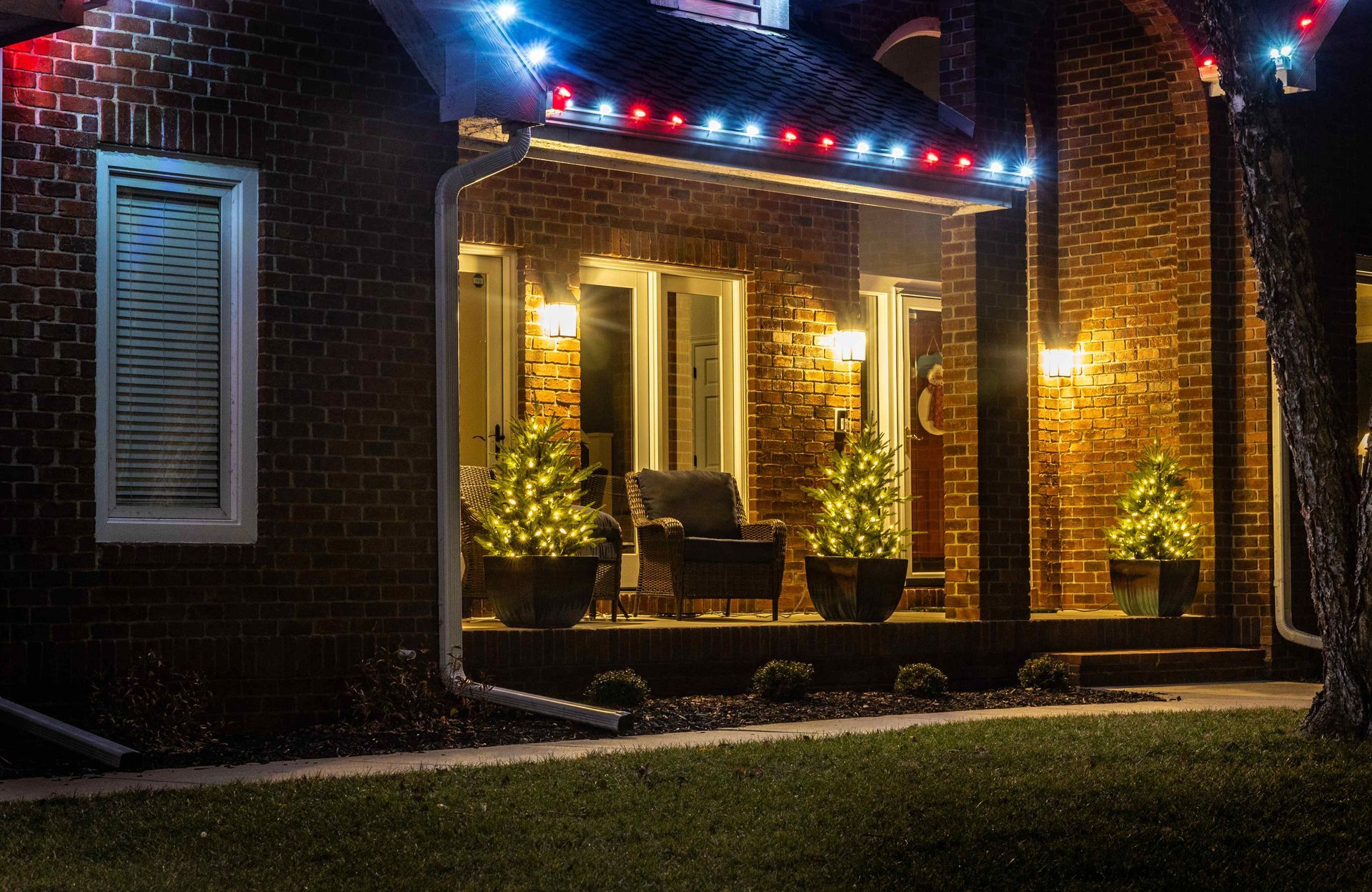 Brick house porch decorated with Christmas lights and small lit trees.
