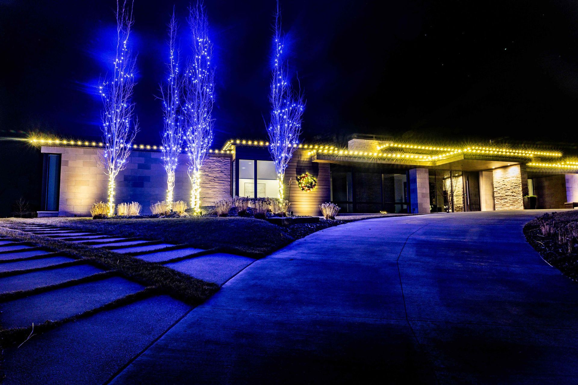 House exterior at night, lit with blue and yellow Christmas lights. Driveway and trees.