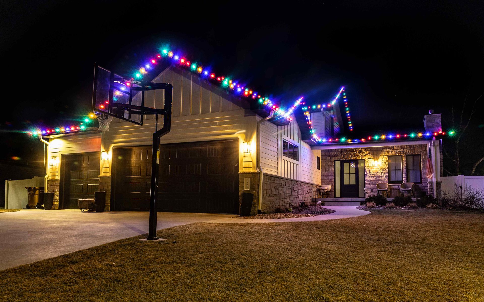 House at night with multicolored Christmas lights along the roof. A basketball hoop is in the front.