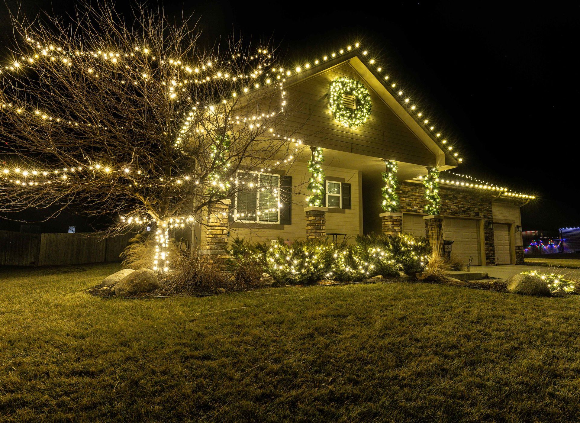 House decorated with warm white Christmas lights, a wreath, and a lit tree, at night.