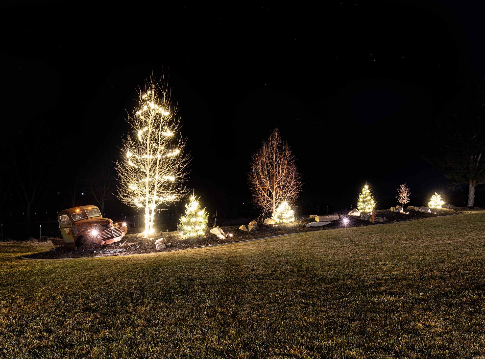Night scene with illuminated trees and a vehicle on a grassy hill; Christmas lights sparkle in the darkness.