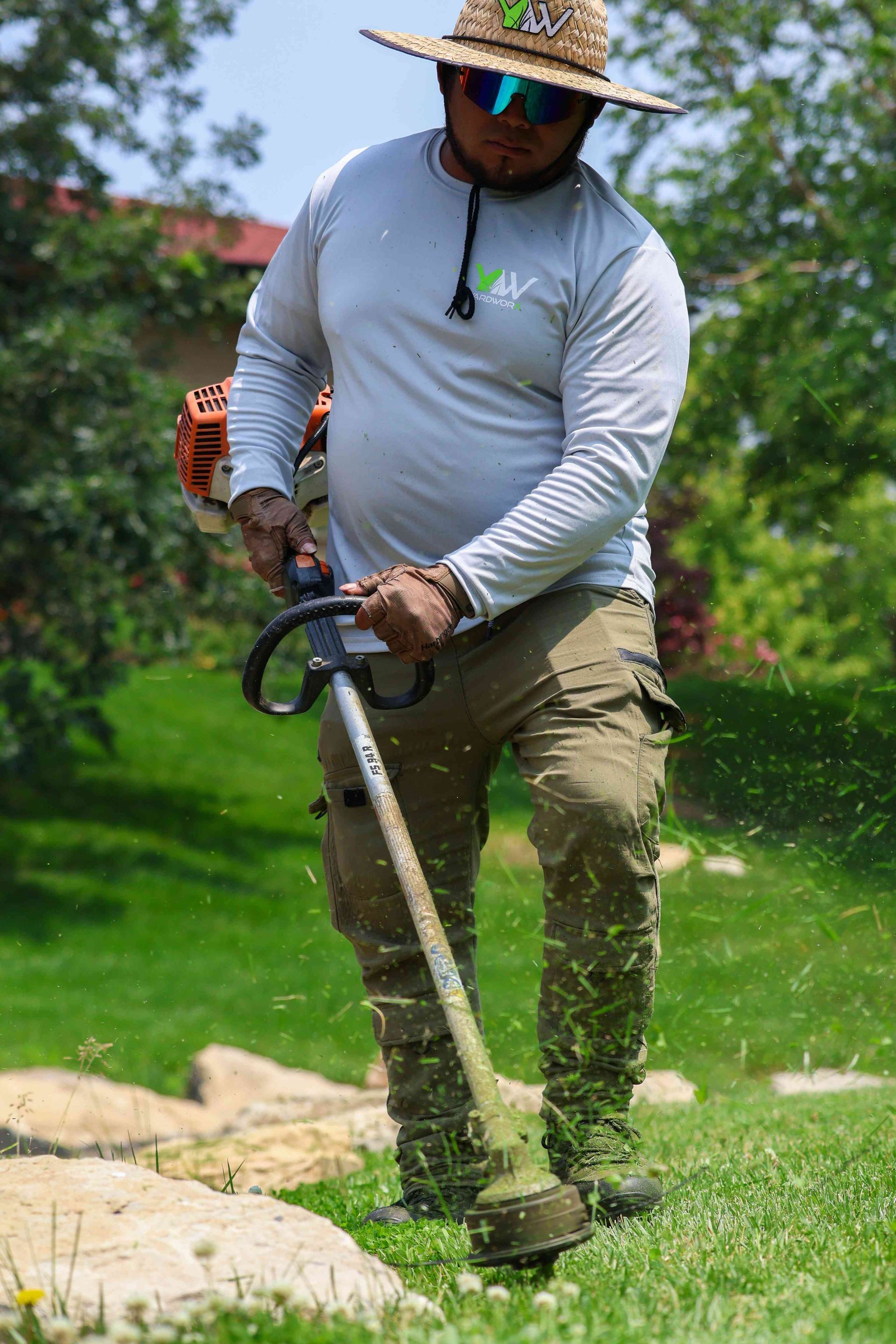 A man wearing a hat and sunglasses is cutting grass with a lawn mower.