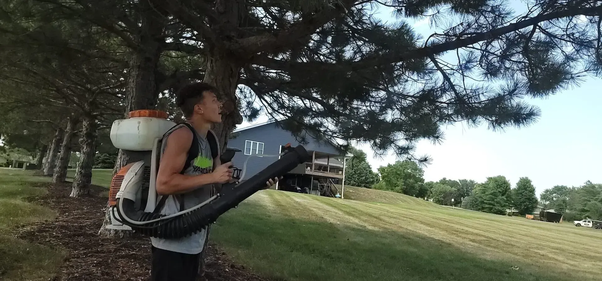 A man with a backpack is standing next to a tree with a house in the background.