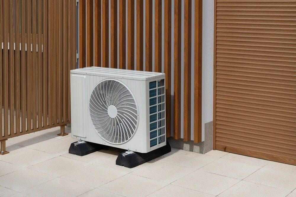 A White Air Conditioner is Sitting on a Tiled Floor Next to a Wooden Fence — AJM's Air Conditioning Centre in Broadwater, NSW