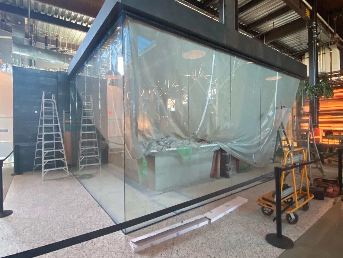 Industrial workspace with a large transparent enclosure, ladders, and construction equipment inside a factory-like setting