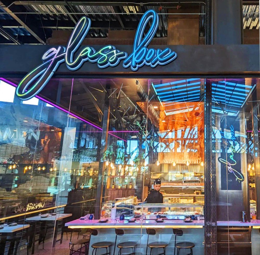 Neon-lit storefront labeled “glass box” with reflective glass windows and colorful interior seating
