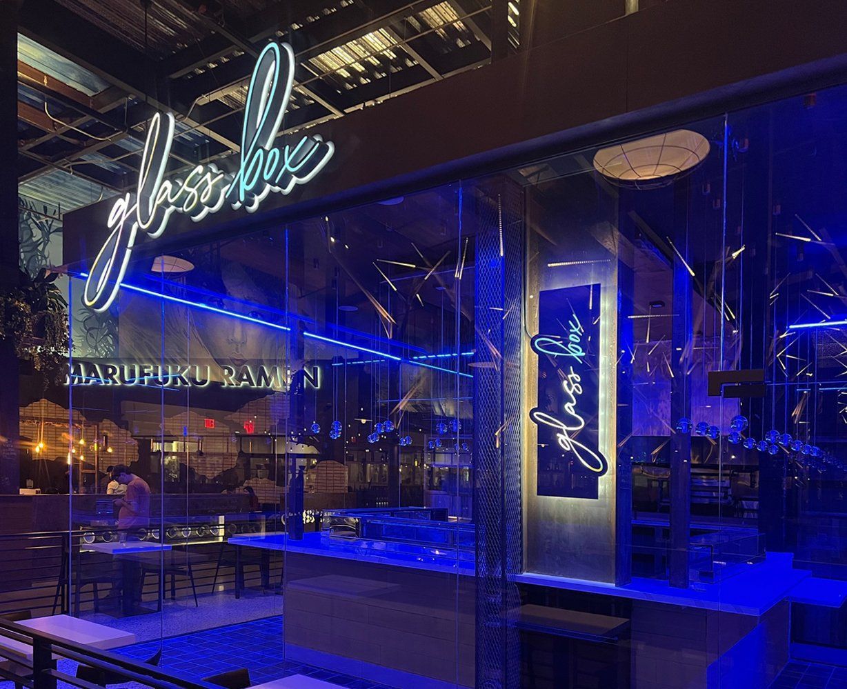 Blue-lit restaurant interior with glass walls, neon sign, and modern tables at night