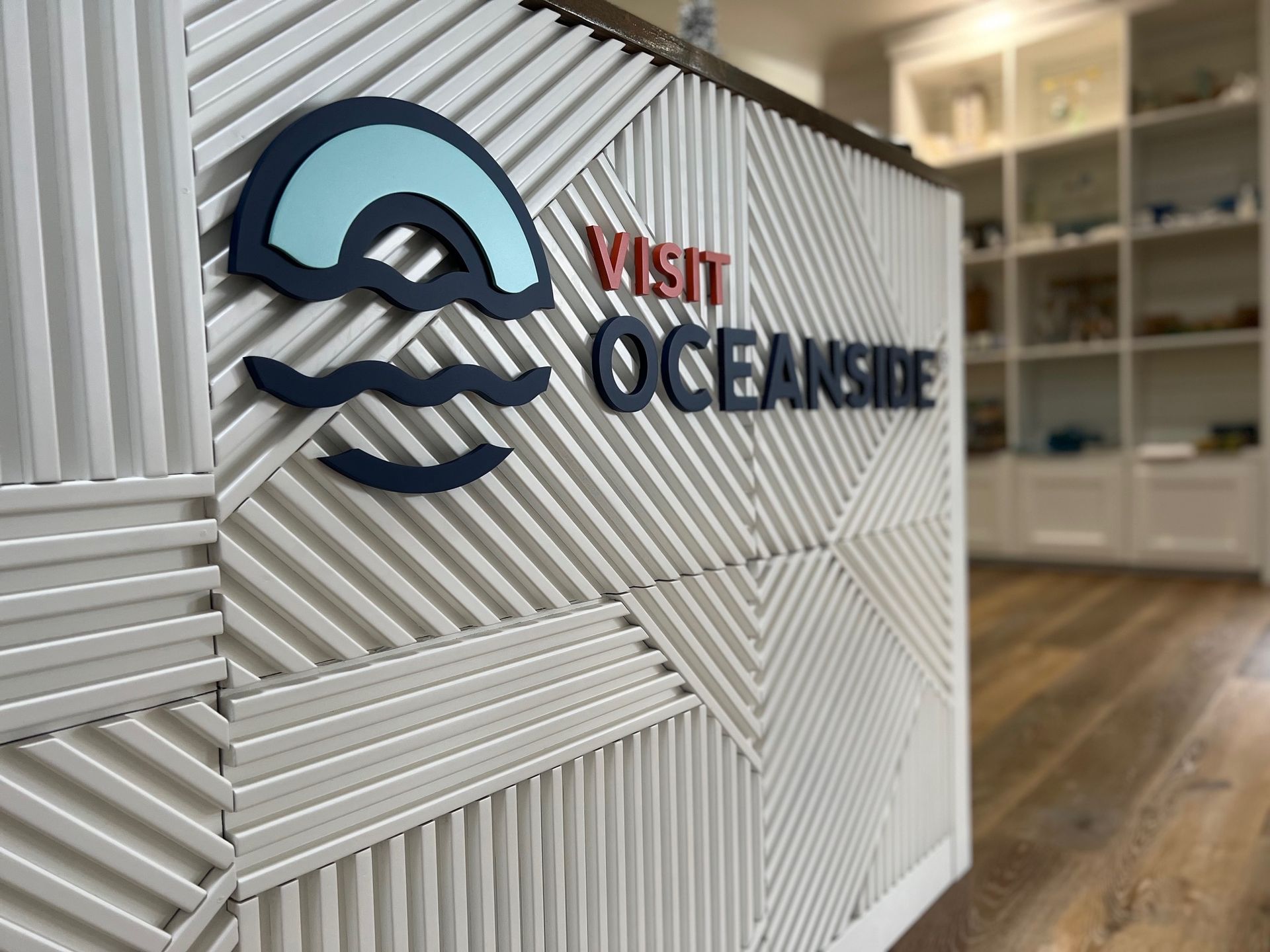 “Visit Oceanside” sign on a white textured wall in a modern office or lobby.
