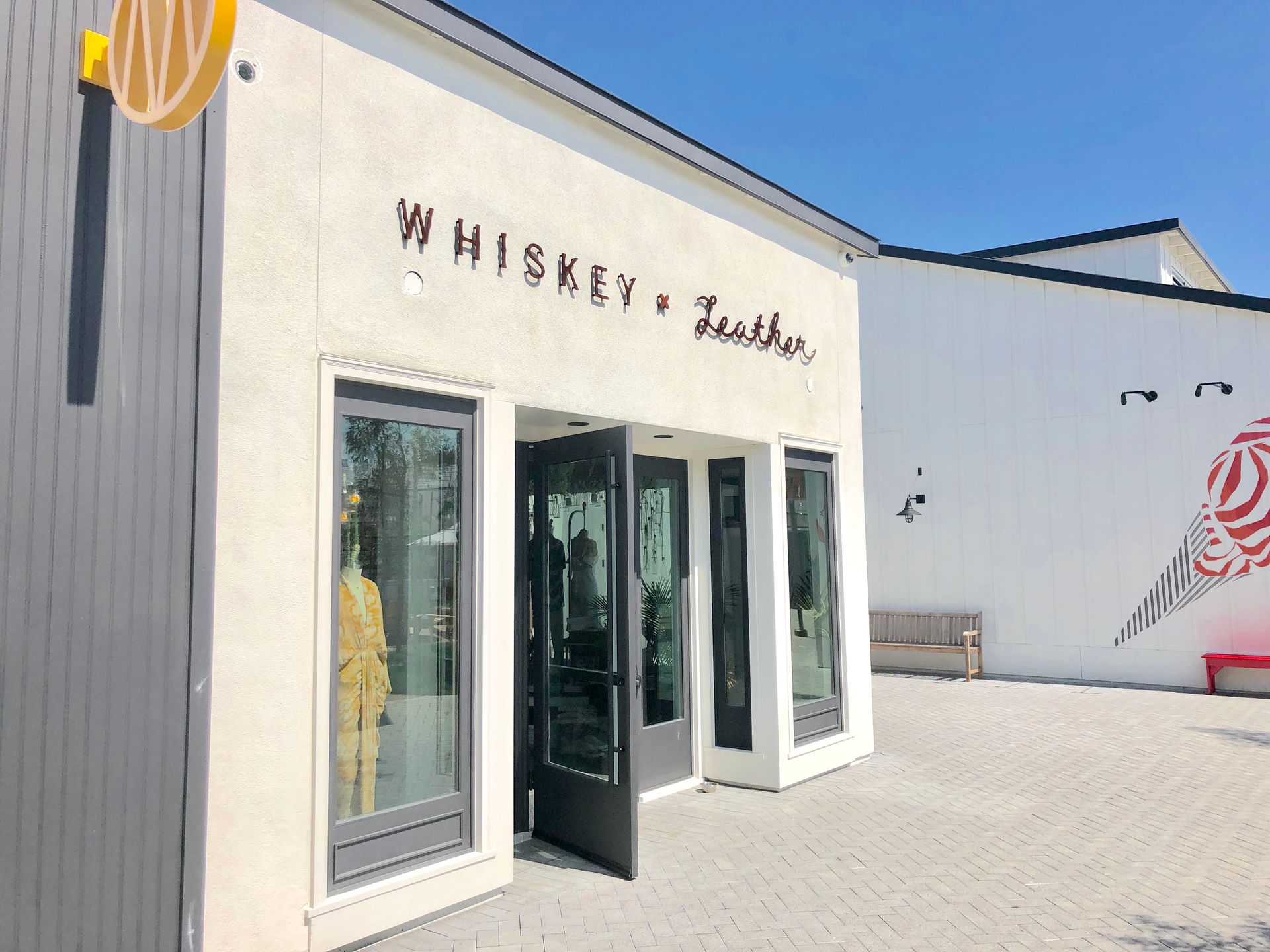 White storefront with open glass doors and “Whiskey” sign, set in a sunny courtyard with blue sky.