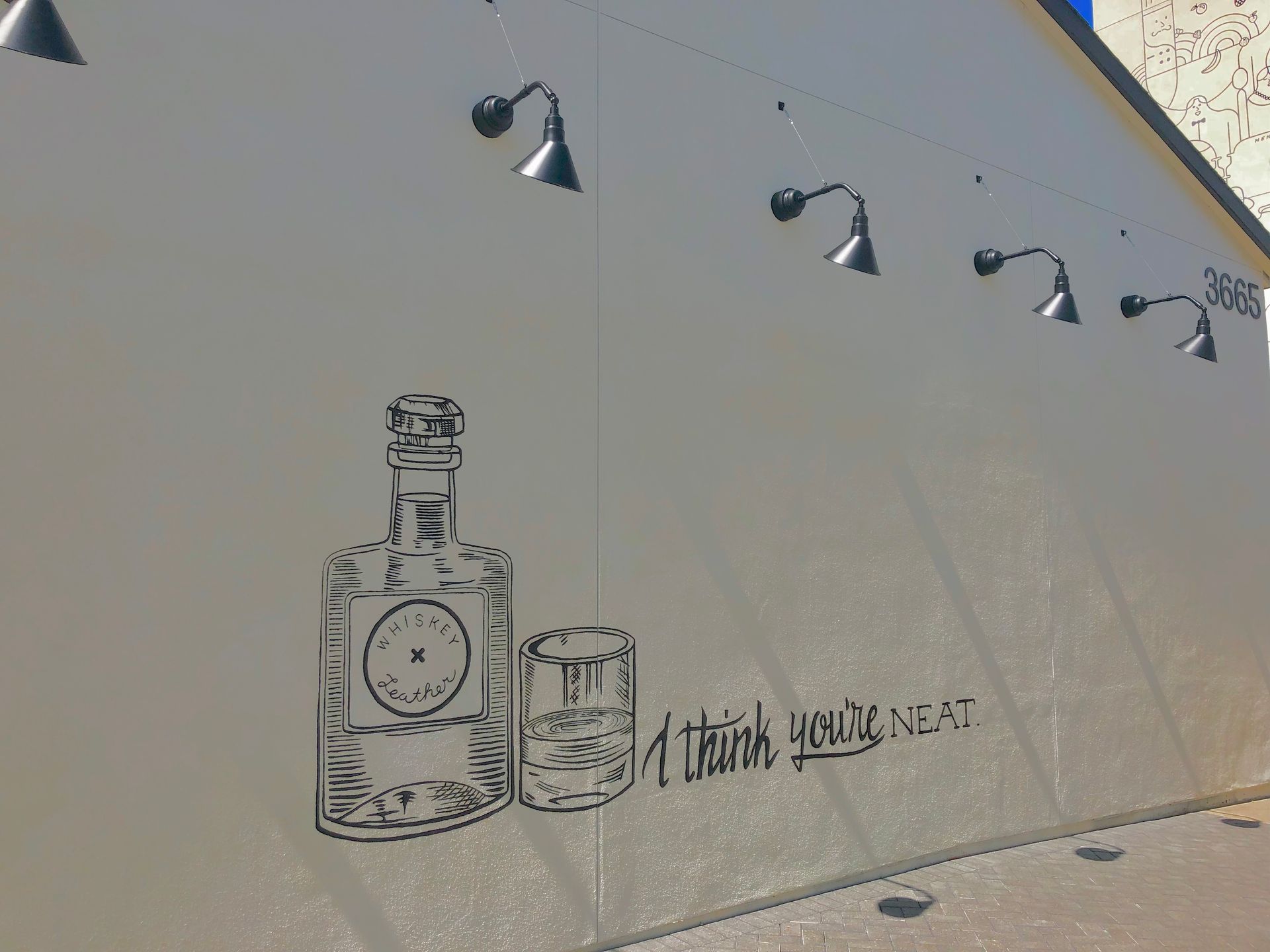 Street mural of a perfume bottle and bottle drawing on a beige wall with small lamps above and graffiti text.