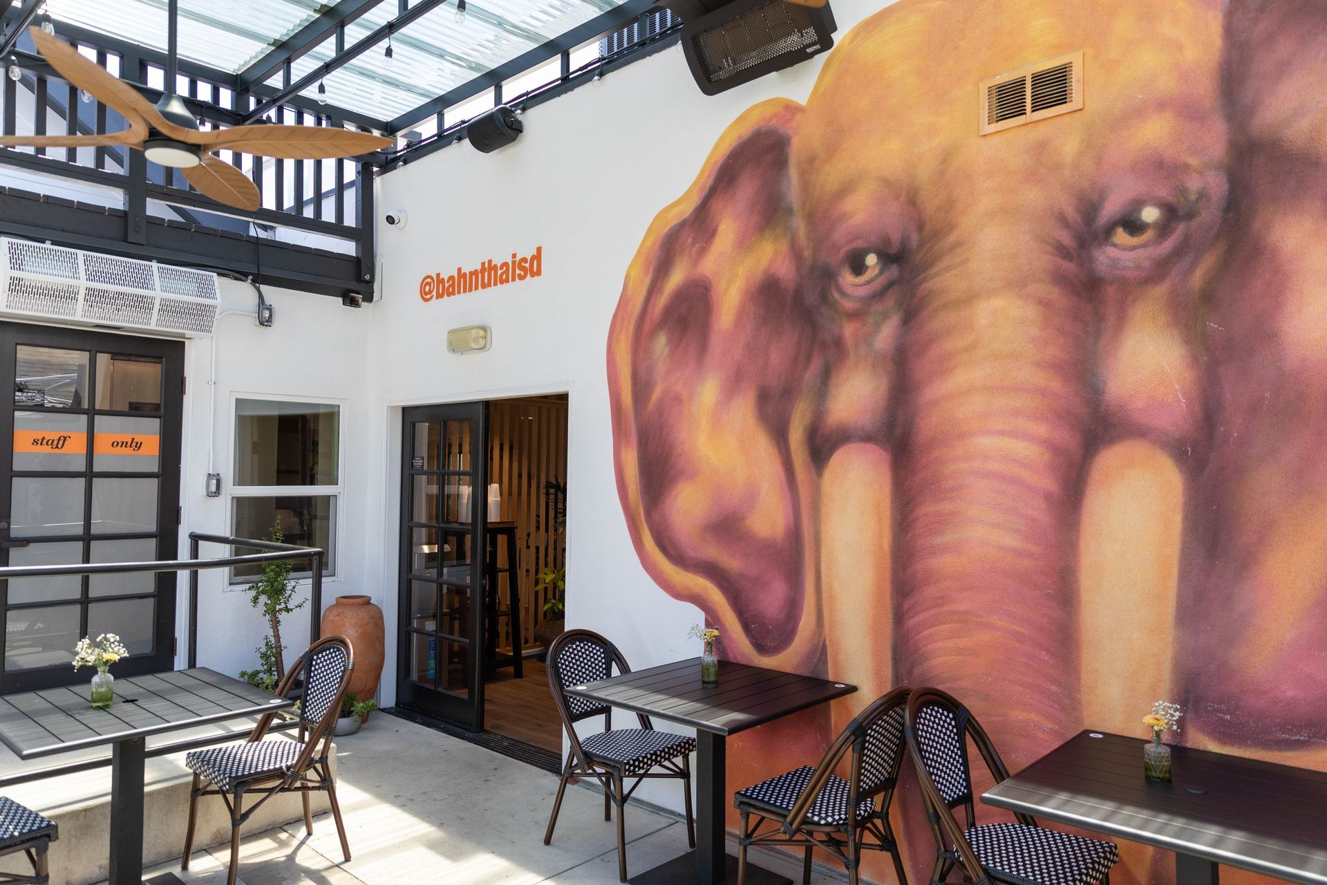 Outdoor café patio with a large elephant mural on a white wall and metal tables and chairs under a glass canopy