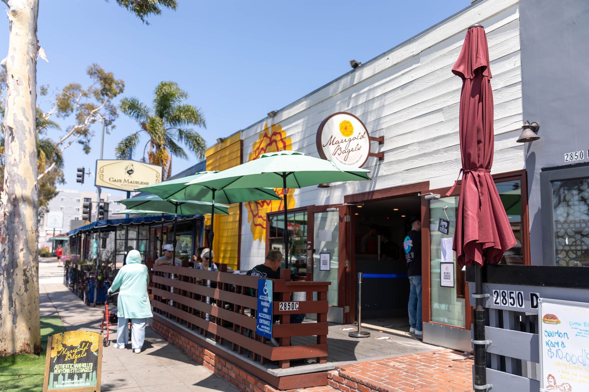 Street-side café with green umbrellas and a person walking past storefronts on a sunny day
