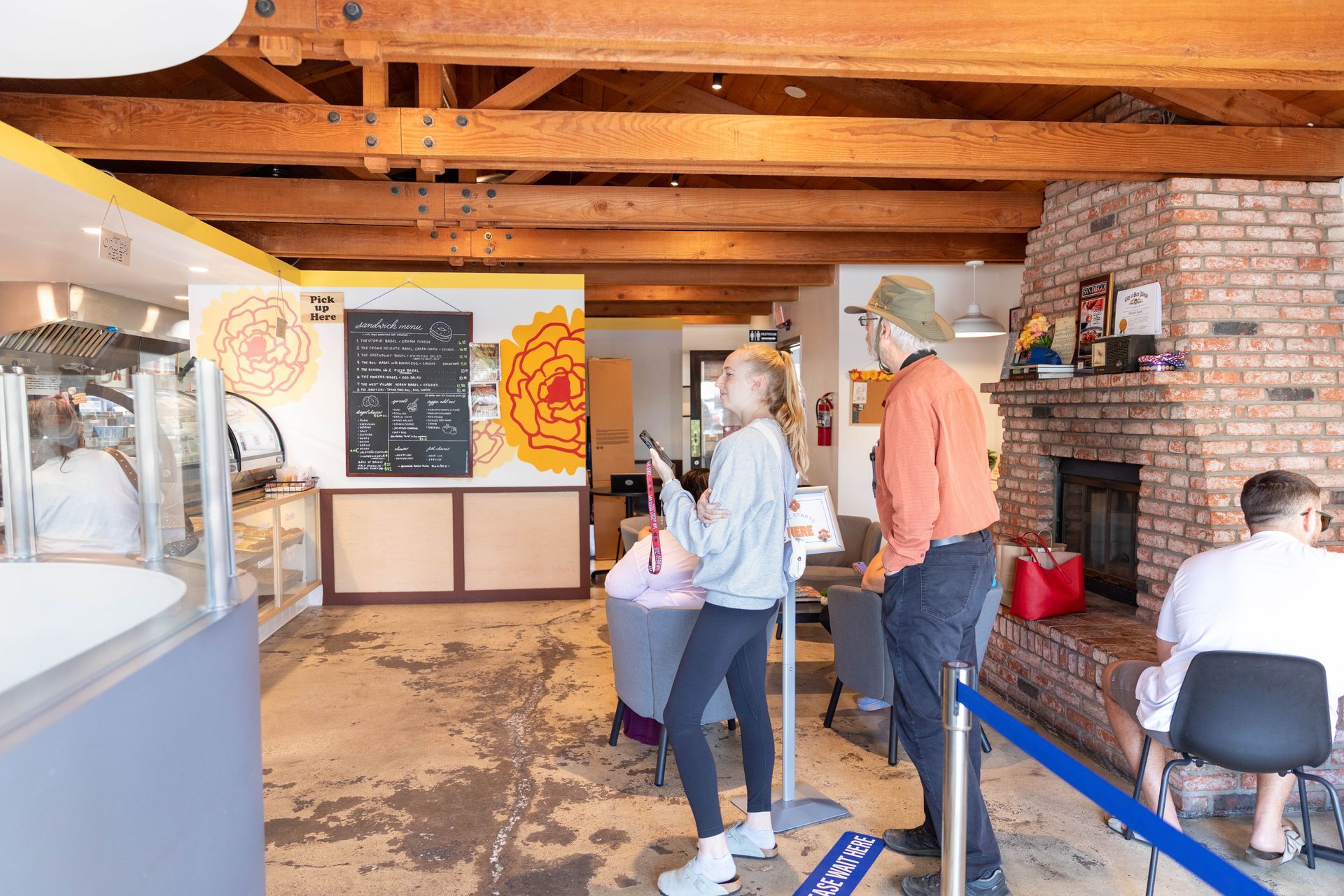 People in a rustic shop, standing by a counter and chalkboard menu near a brick fireplace.
