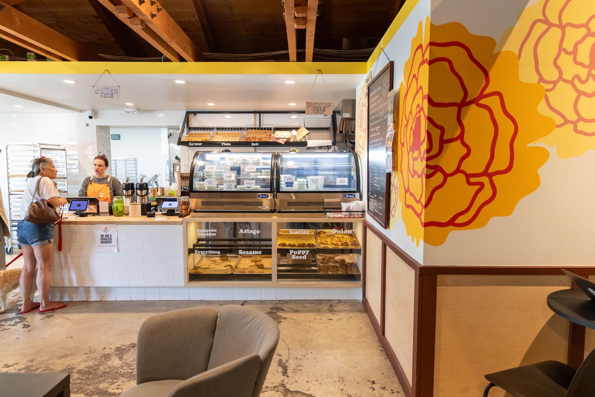 Bright café interior with pastry display case, counter staff, and a large floral wall mural