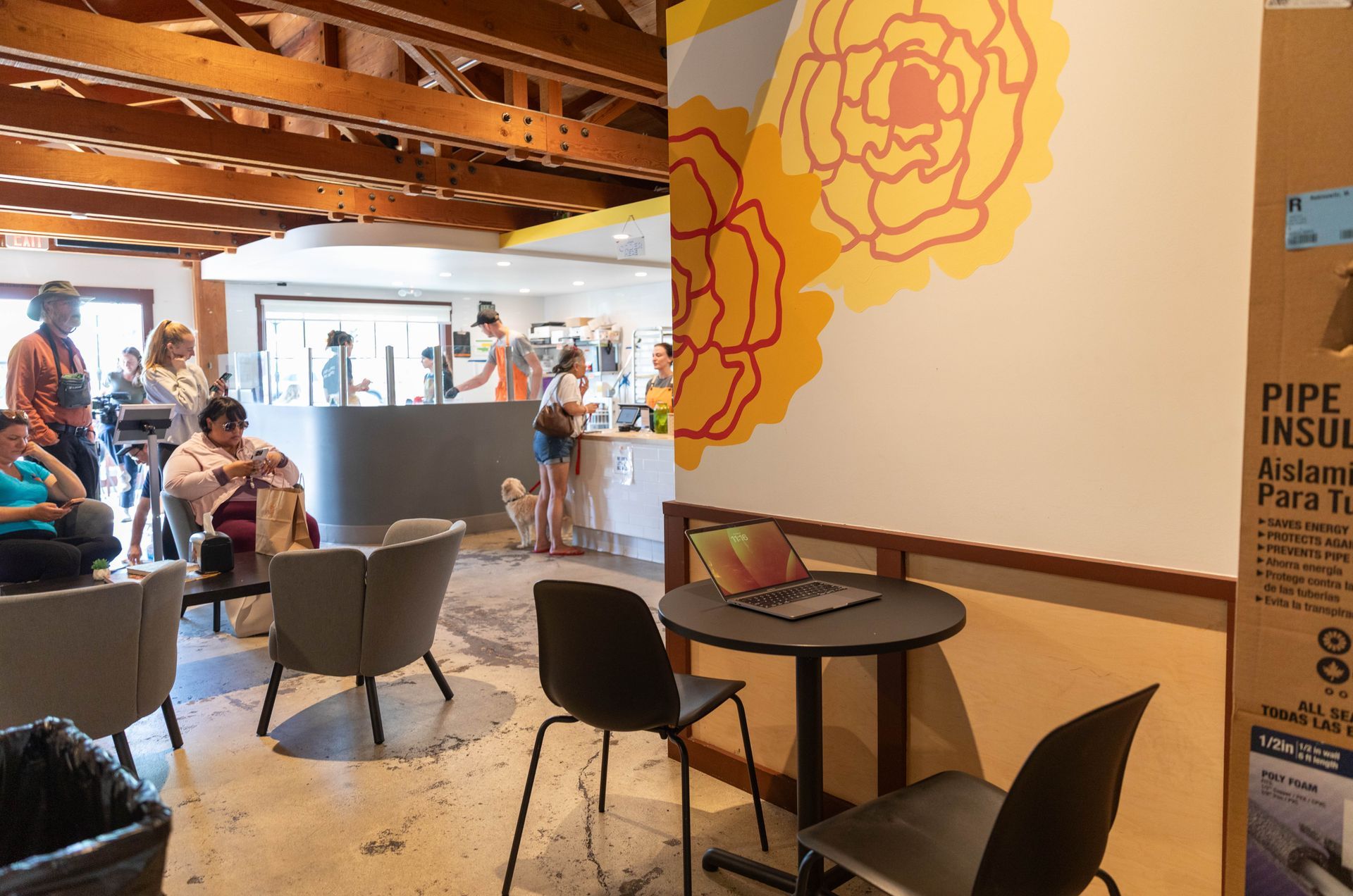 Modern cafe lounge with people seated at tables under wooden beams and a floral wall mural