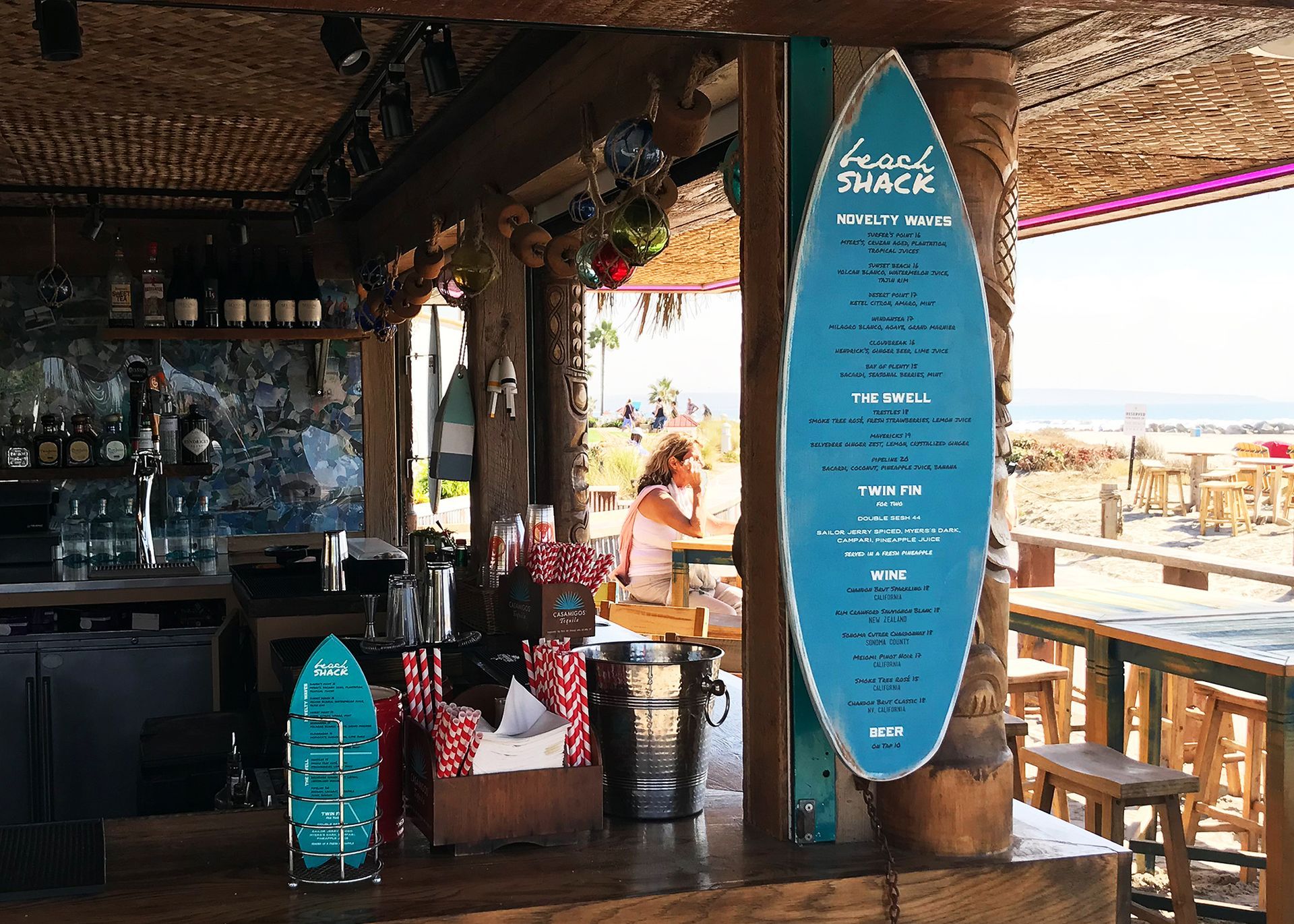 Beach bar interior with surfboard-shaped blue menu, wooden stools, and ocean view beyond open deck.