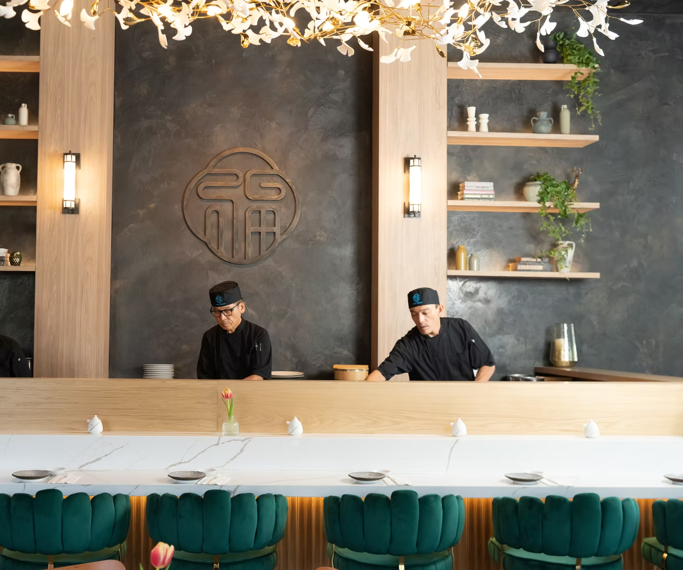 Modern restaurant interior with two chefs behind a long counter and teal chairs in front