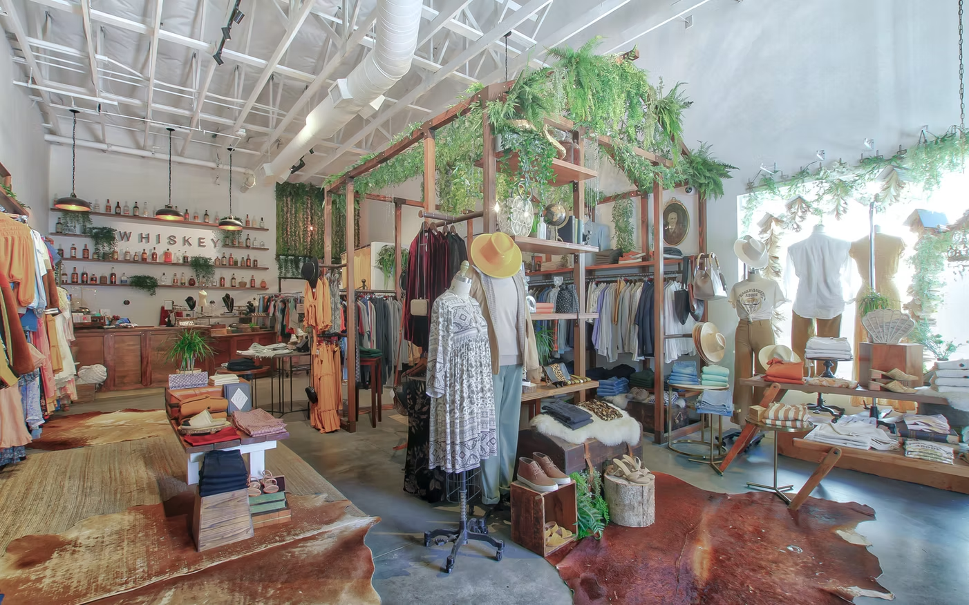 Bright clothing boutique with dresses, scarves, shelves, and decorative plants under a high white ceiling
