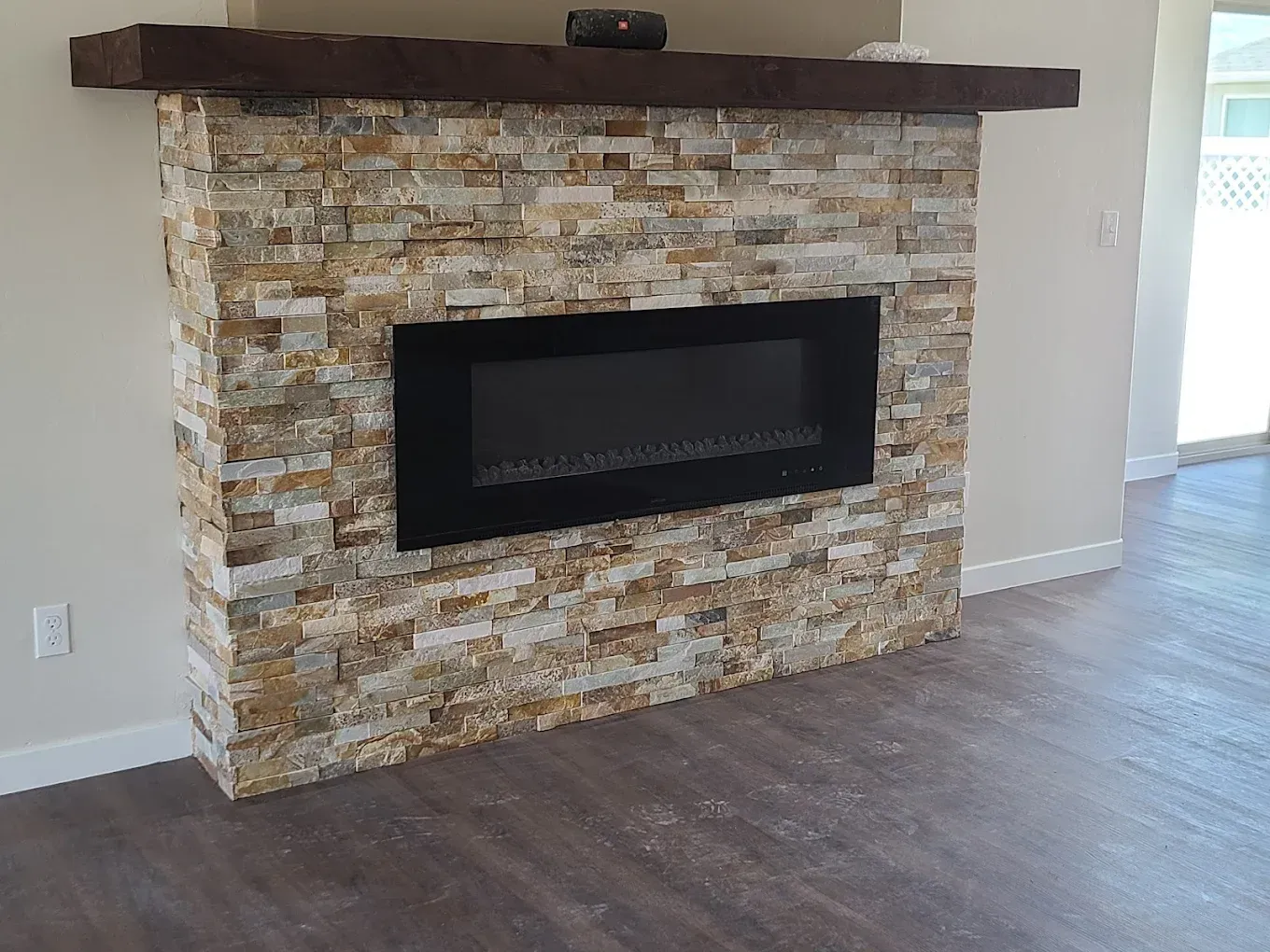 Fireplace with stone facade, black electric insert, and wooden mantel. Set in a room with hardwood flooring.