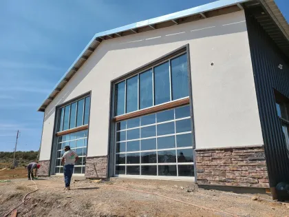 Building exterior with large glass garage-style doors, white stucco, stone base, workers, and a blue sky.