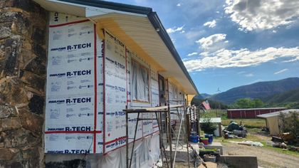 House exterior under renovation; stone wall, R-Tech insulation, scaffolding, blue sky, mountains in the background.