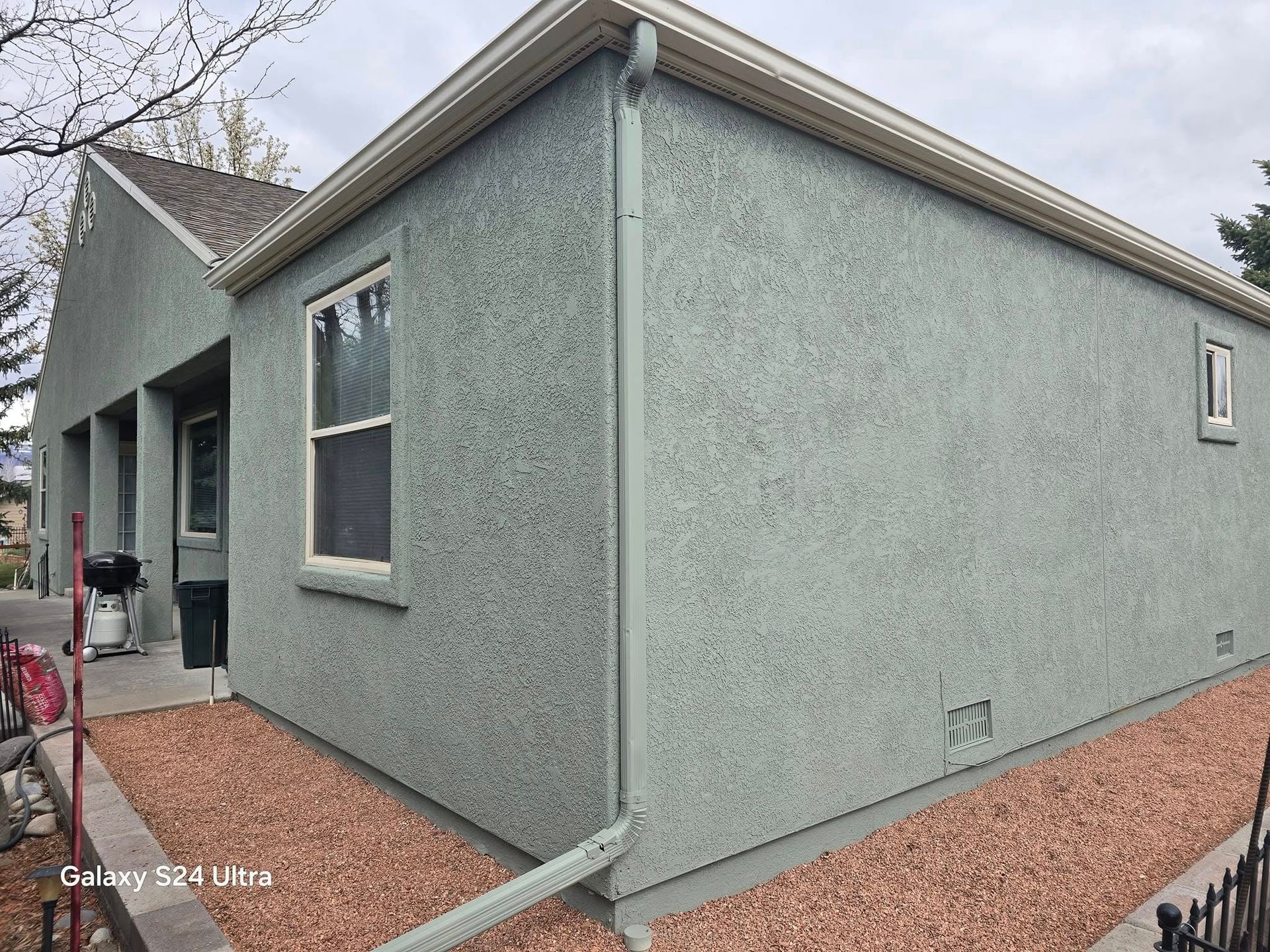 Exterior of a light green stucco house with gravel landscaping, a window, and a gutter.