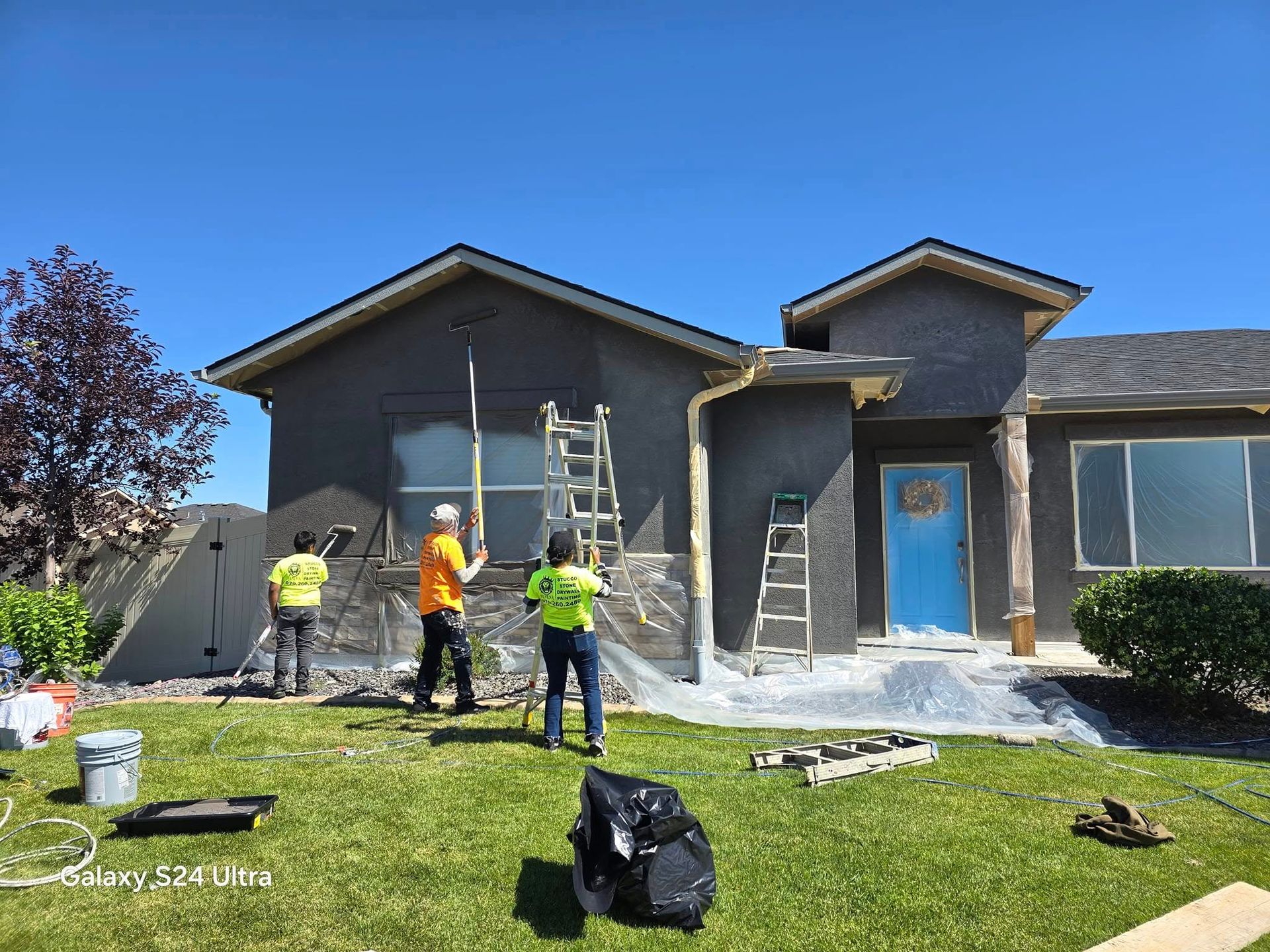 Three workers applying stucco to a house exterior; a light blue door, ladders, and a green lawn.