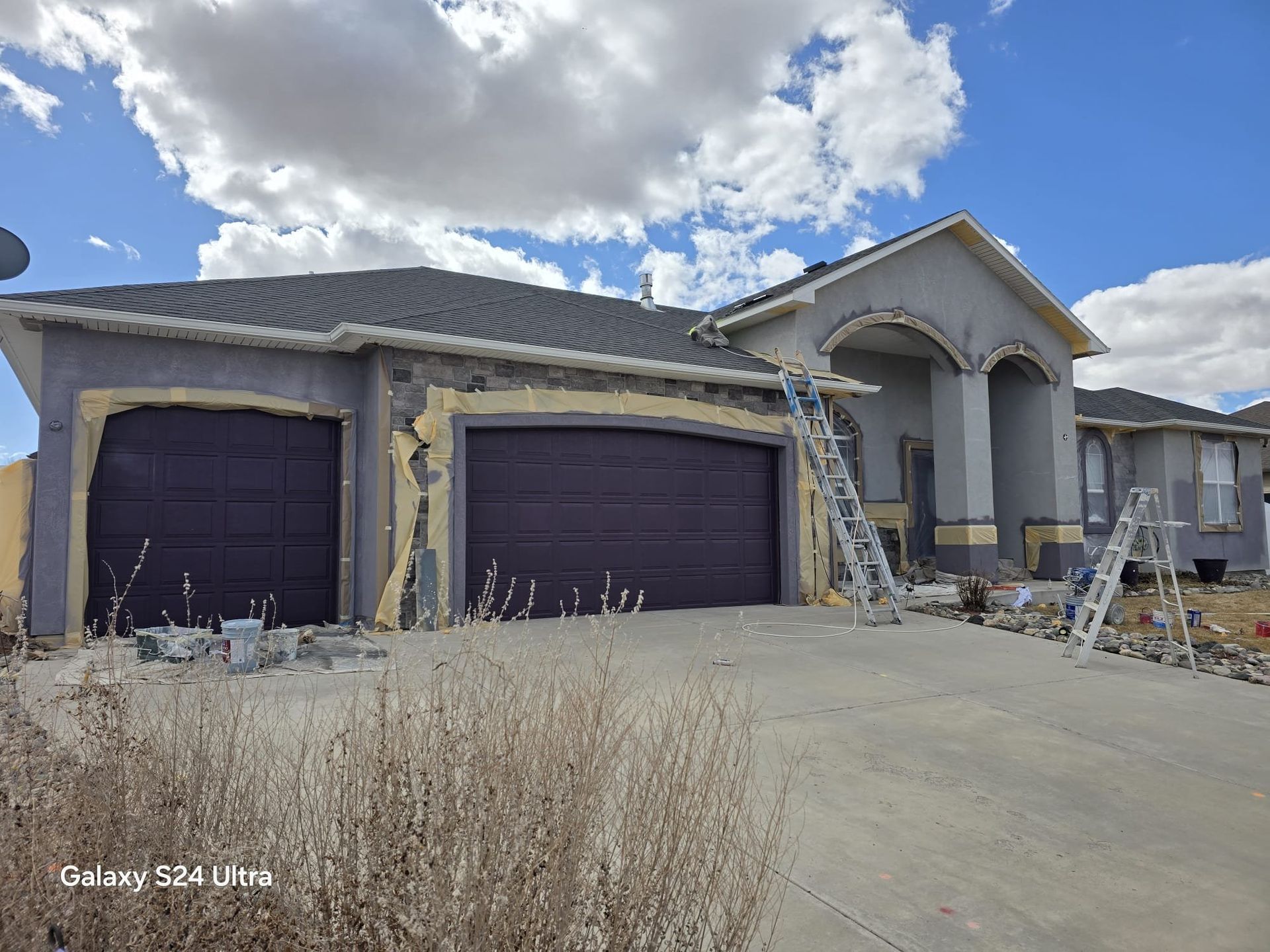 House exterior under construction with grey stucco, purple garage doors, ladders, and a cloudy sky.
