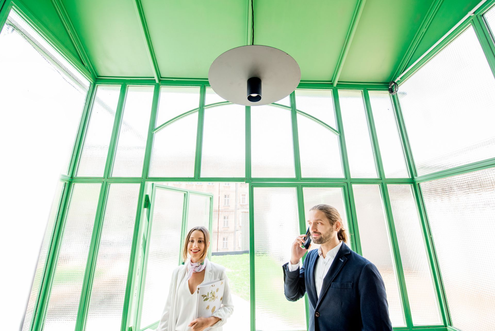 A man and a woman are standing in a green room talking on cell phones.