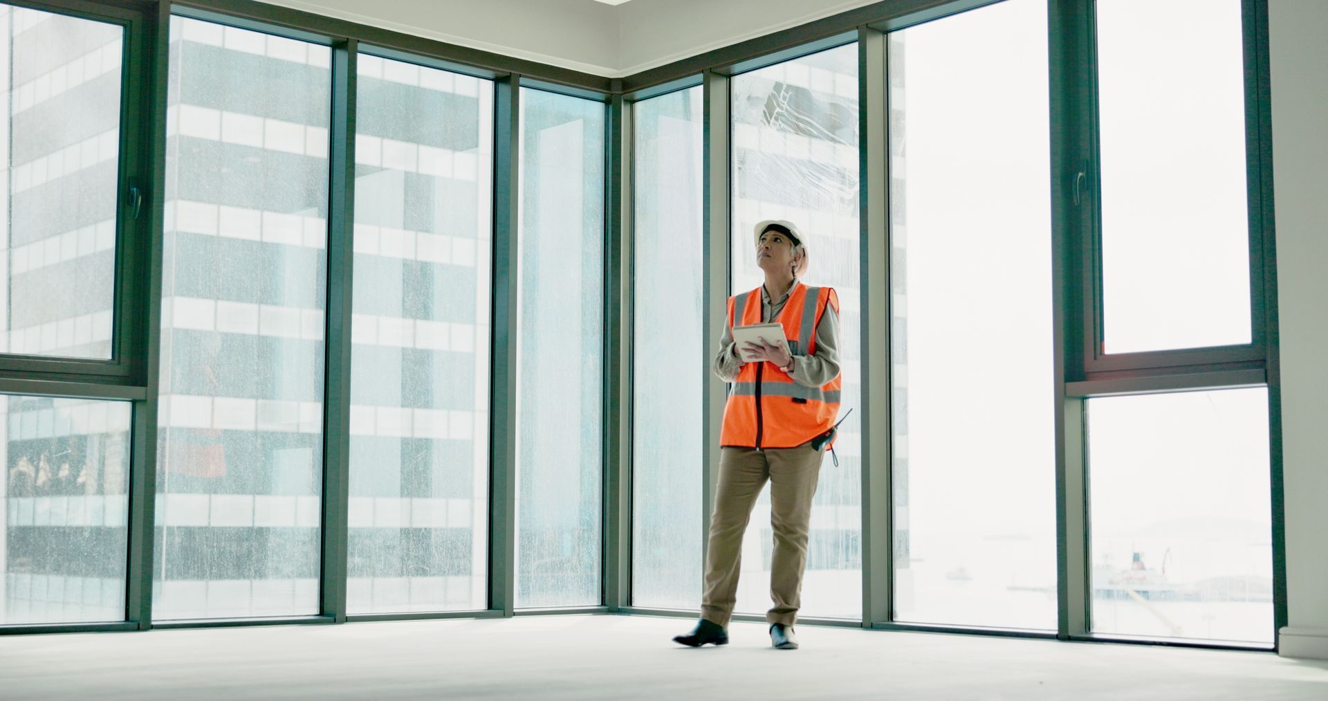 A man in an orange vest is standing in an empty room with a lot of windows.