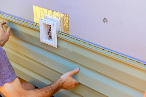Hands installing green vinyl siding near a white vent and wires.