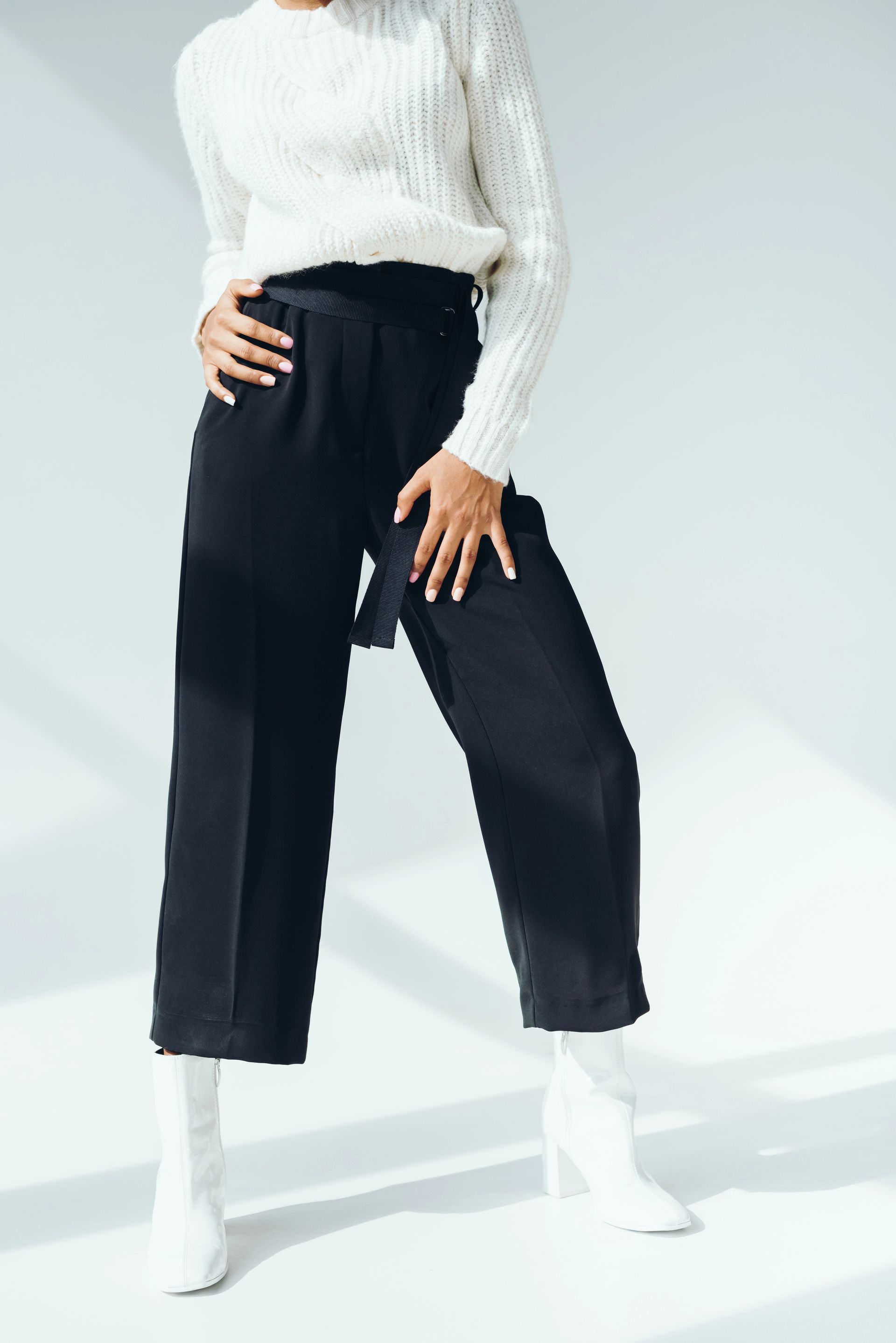Mujer con suéter blanco, pantalón negro y botas blancas. De pie sobre fondo blanco, con las manos en la cintura.