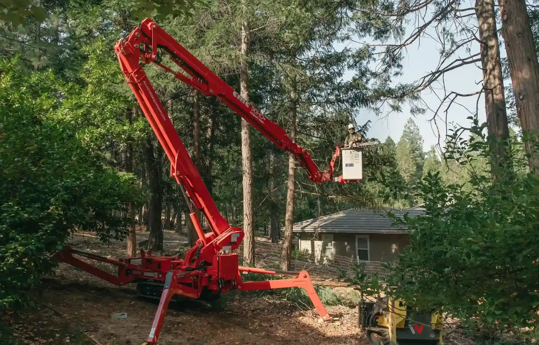 Tree limb being lifted by a crane over a suburban house, with debris piled on the driveway.