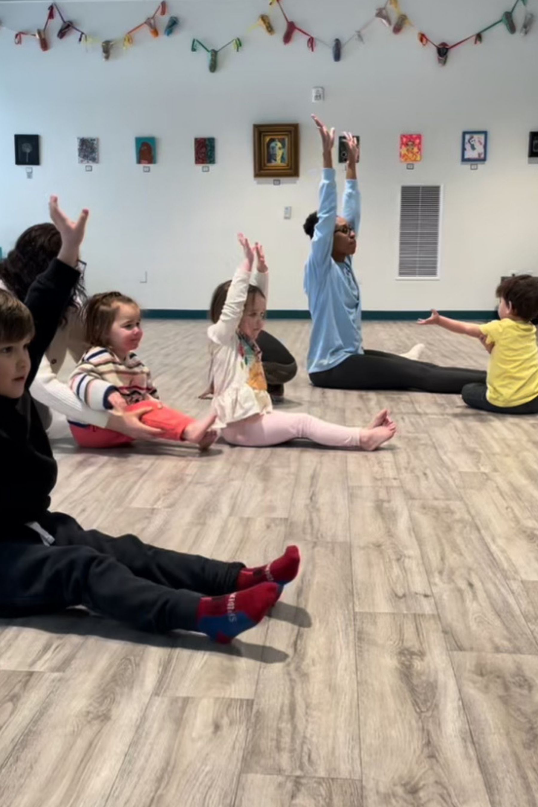 Children in a room with a teacher, seated with legs extended, arms raised.