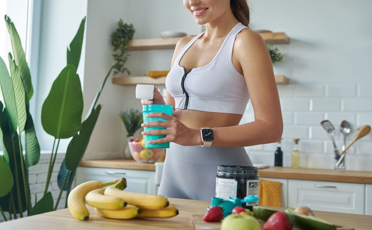 Mujer en ropa deportiva preparando un batido en una cocina con frutas y suplementos.