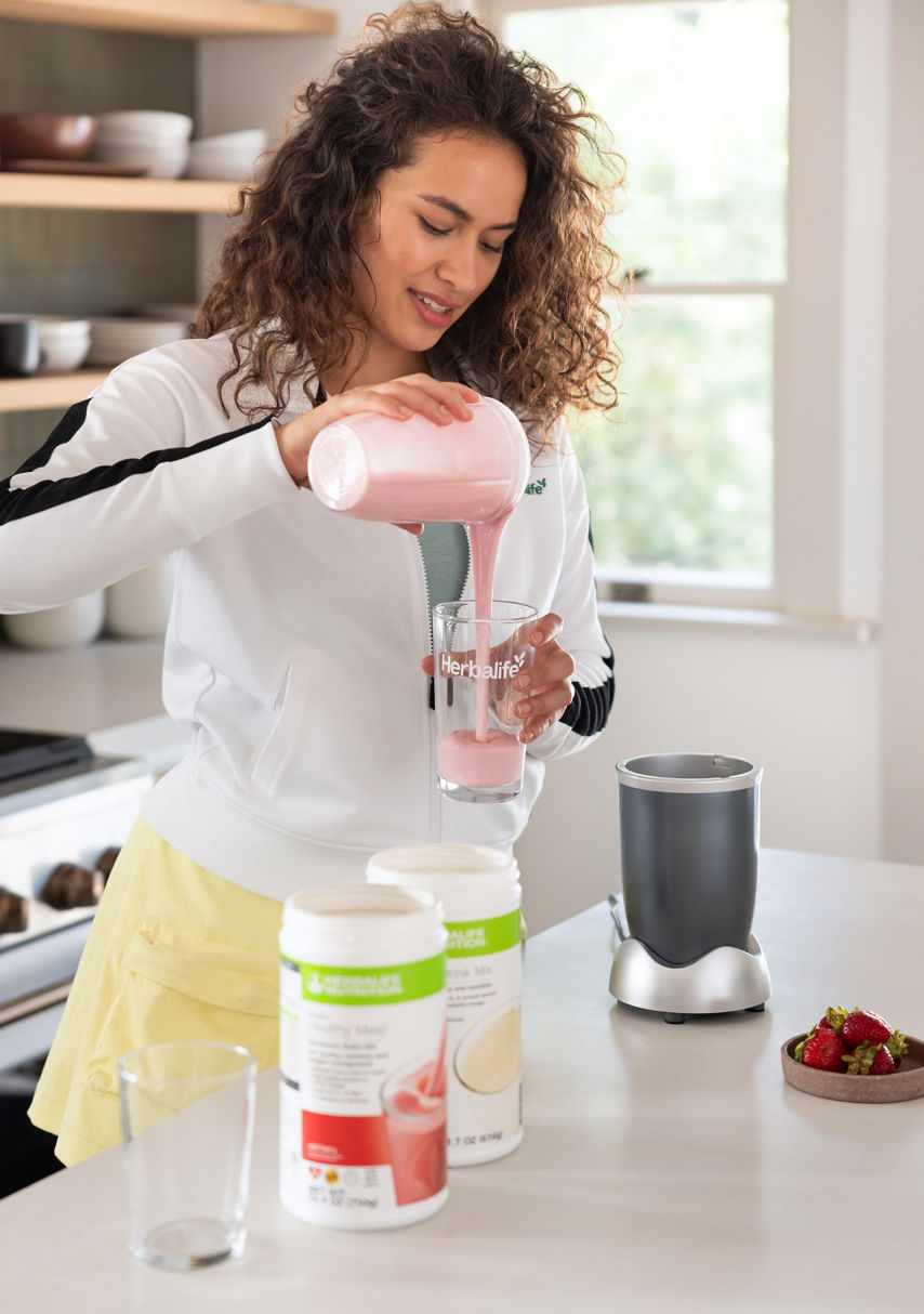 Mujer vertiendo batido rosa en un vaso en la cocina, licuadora y frascos cerca.