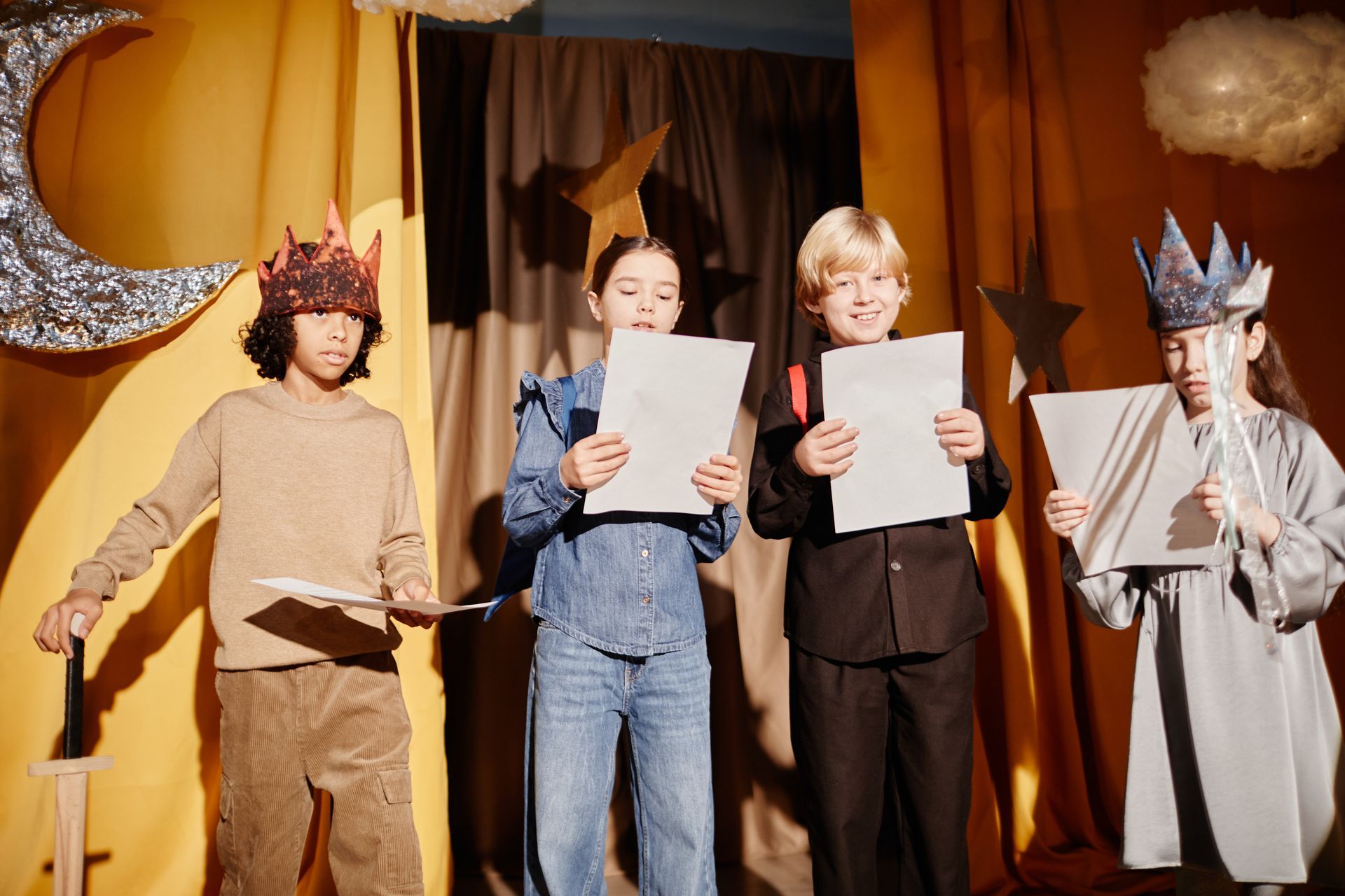 Children in costumes on a stage hold papers, reading. A crescent moon and stars decorate the backdrop.