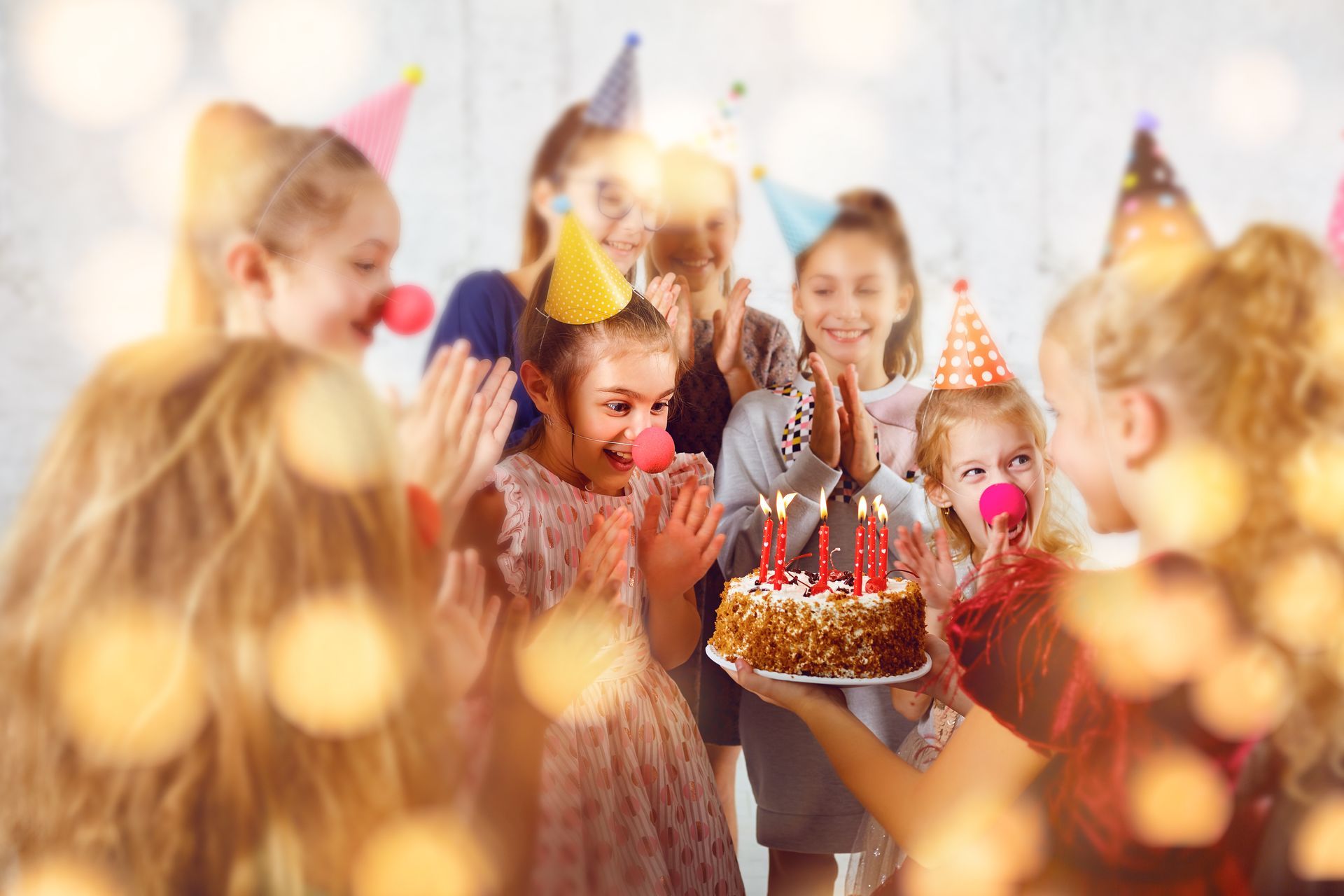 Children at a birthday party, gathered around a cake with lit candles. They are clapping and smiling.