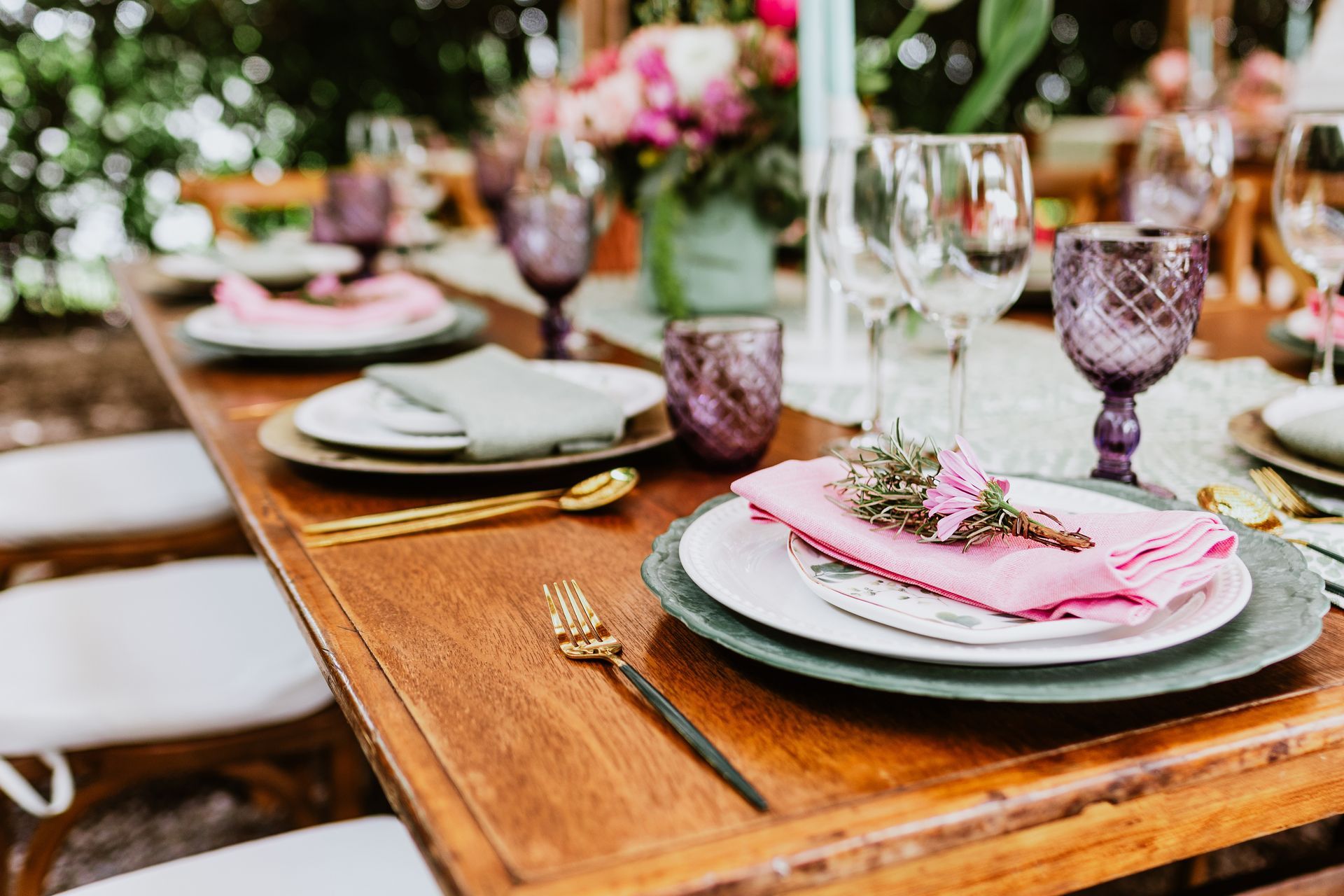 Wooden table set for a formal outdoor meal with pink napkins, gold flatware, and purple glassware.