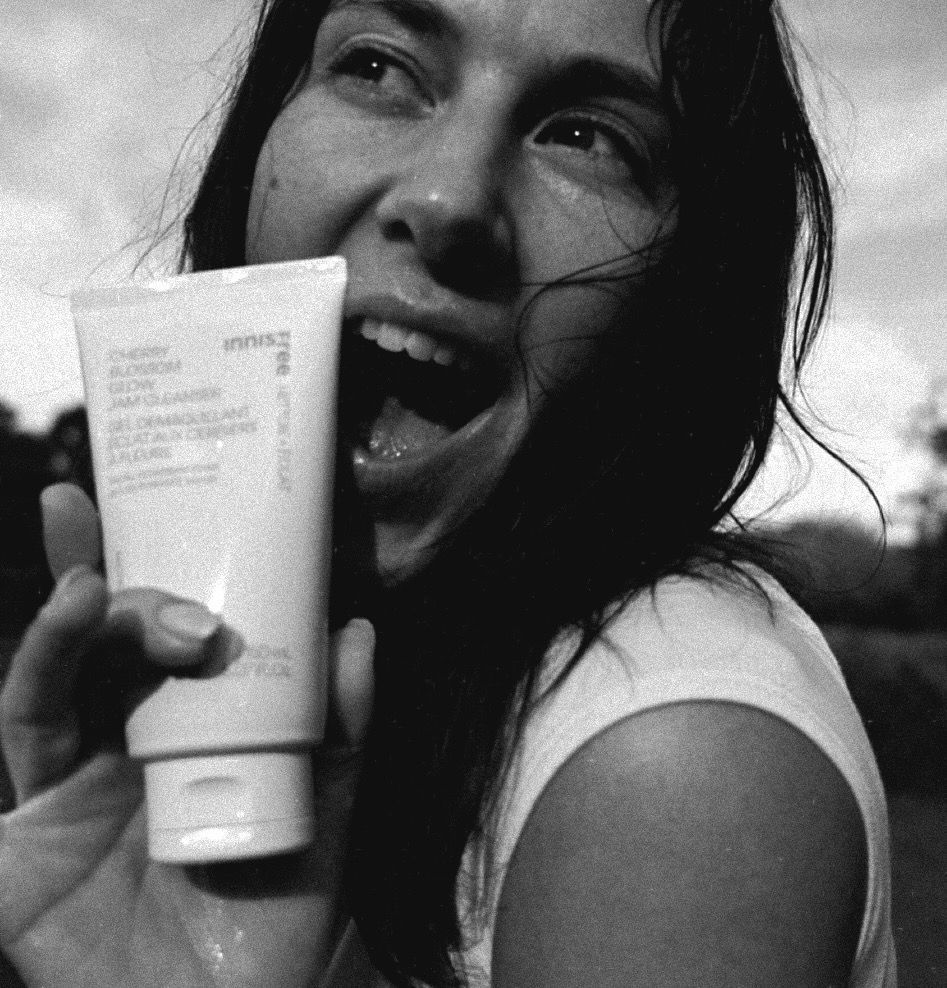 Woman excitedly holding a tube of skincare product, smiling and looking up, outdoors.