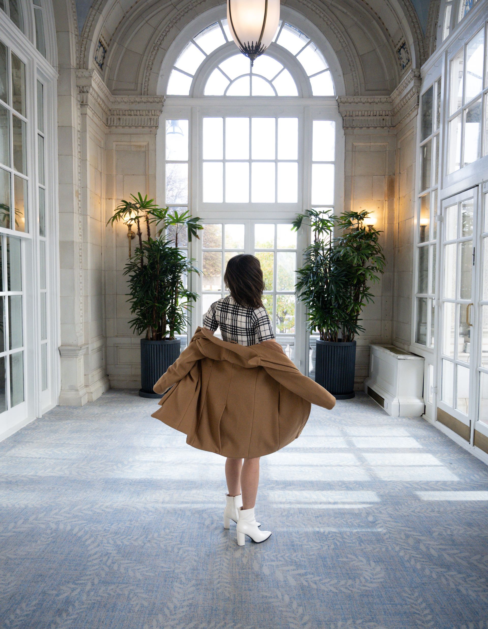 Woman in dress and coat in a grand hallway with large windows and potted plants. She is turning with coat open.