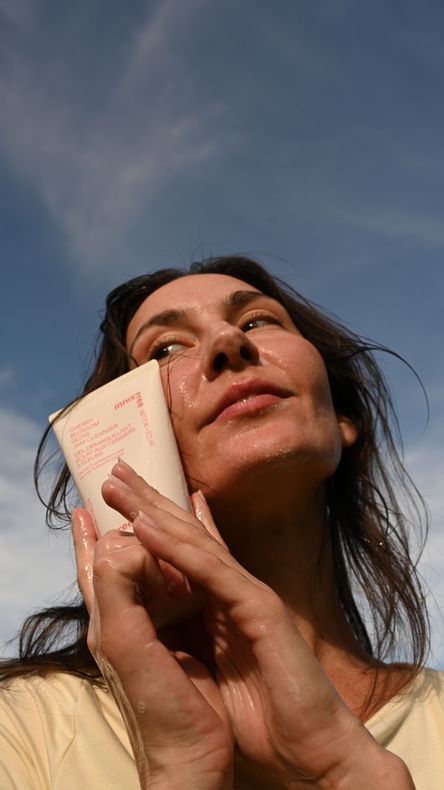 Woman looking up, holding a tube to her face, with a sky background. She's outside and appears to be applying product.